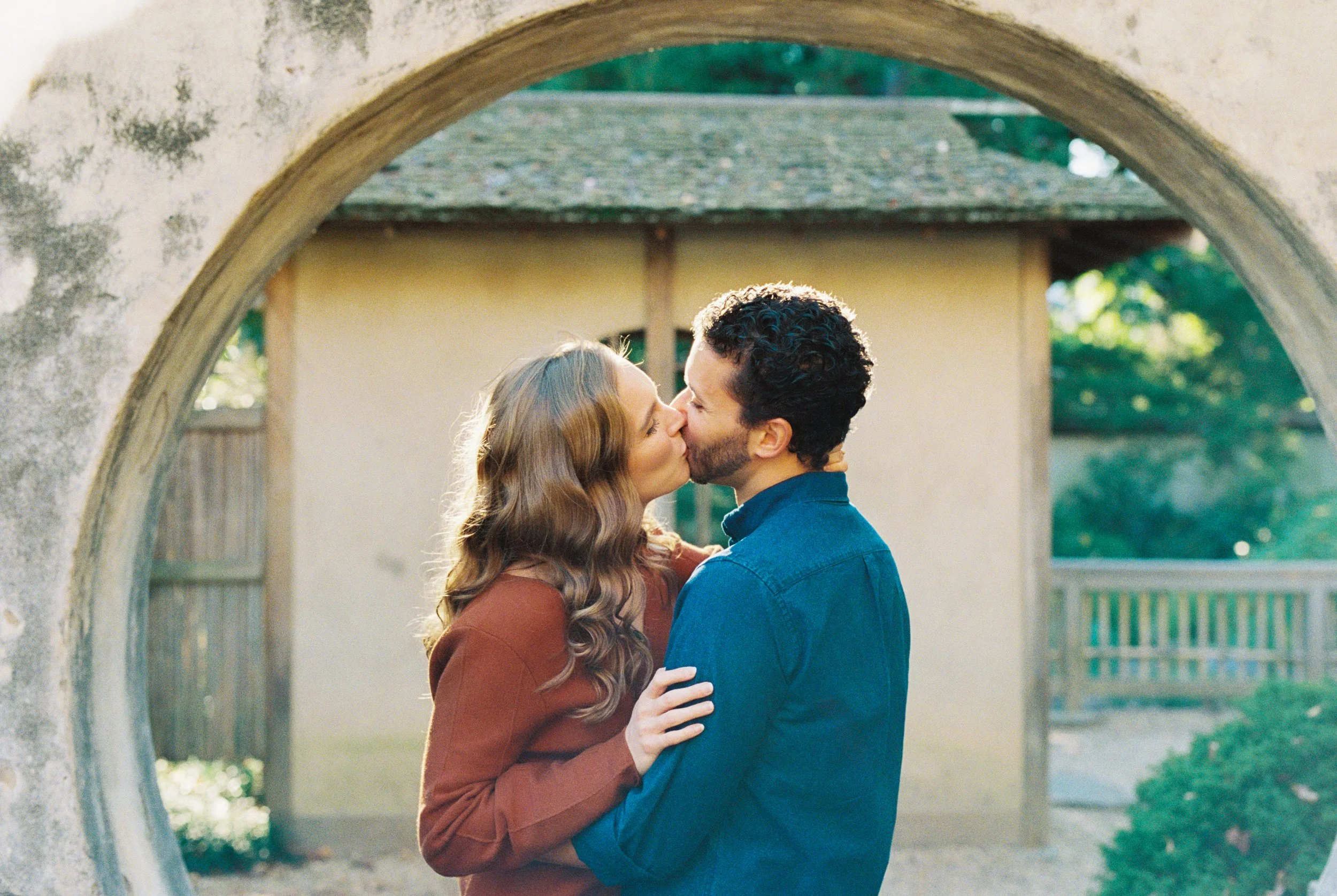 Couple kissing beneath a round stone archway near the garden buildings