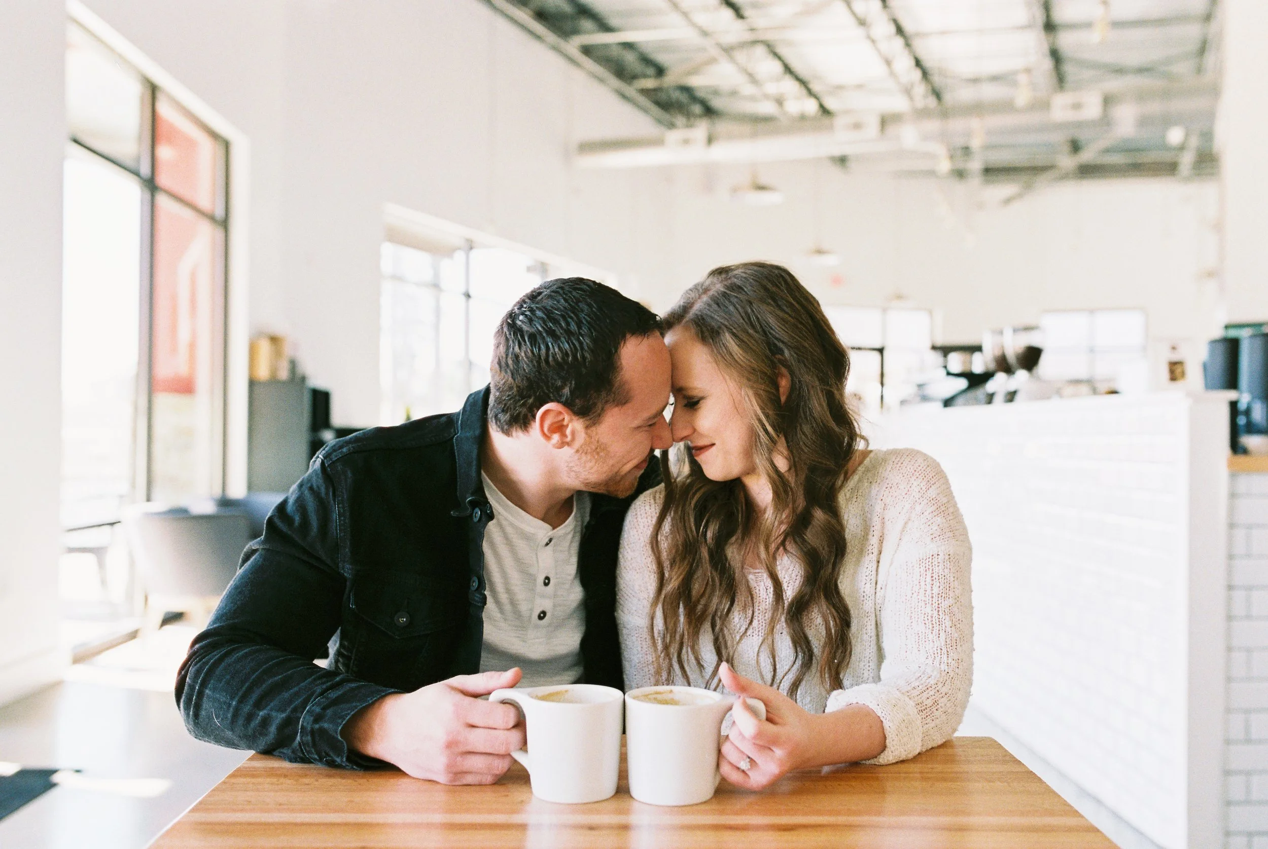 Conner and Maddie sit forehead to forehead at a wooden table with coffee mugs in a Dallas, Georgia coffee shop.