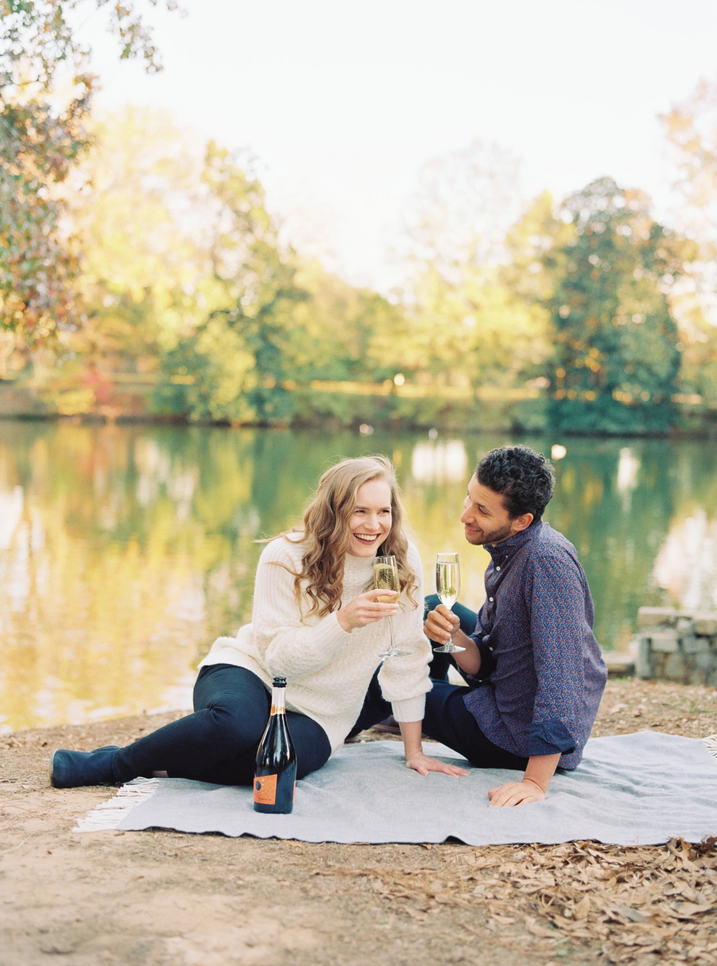 Couple laughing together while toasting champagne on a picnic blanket at the lake's edge