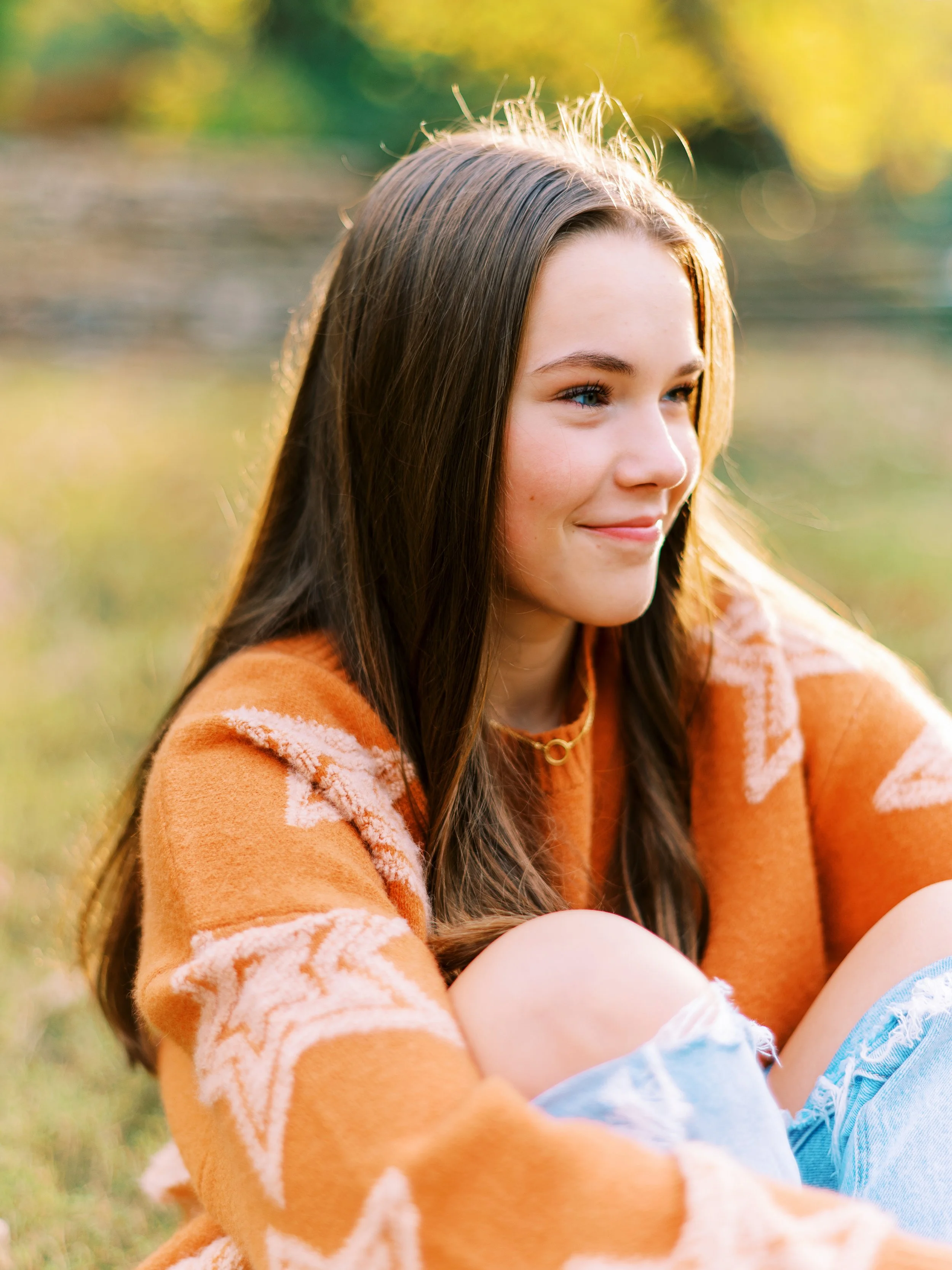 Senior portrait of Hannah sitting and hugging her knees while looking off camera, wearing an orange star-patterned sweater at Kennesaw Mountain Battlefield Park in Kennesaw, Georgia