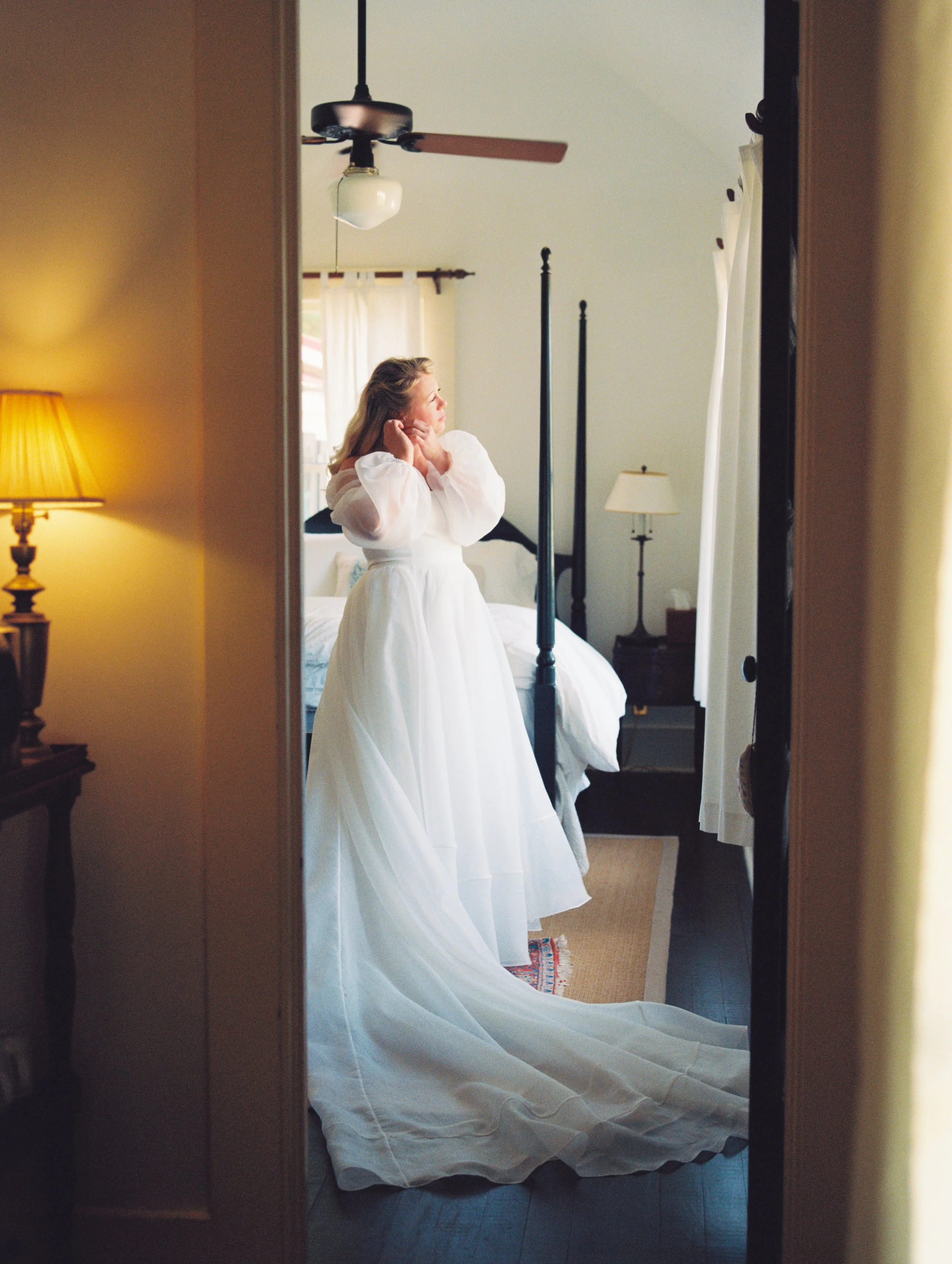 A woman in a southern white wedding dress standing in front of a mirror adjusting her hair, viewed through a doorway in a bedroom with a bed, lamps, and ceiling fan at The Greyfield Inn on Cumberland Island.