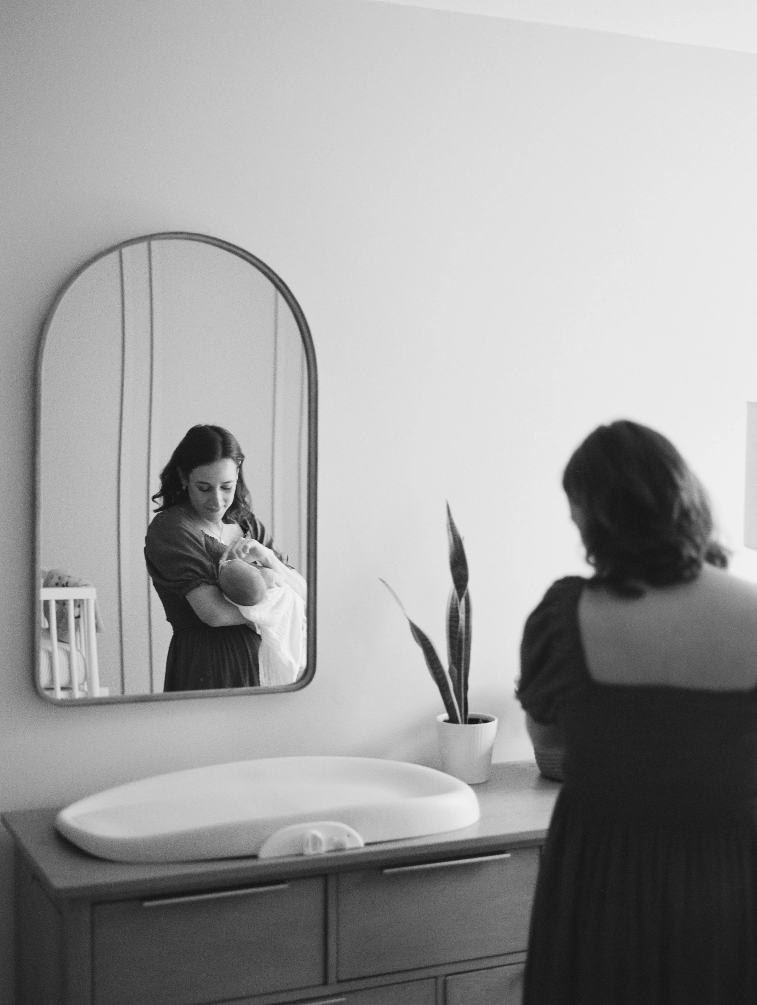 A woman holding a baby, seen in a mirror, standing next to a dresser with a potted plant and a changing pad. Both women are in a room with minimal decor.