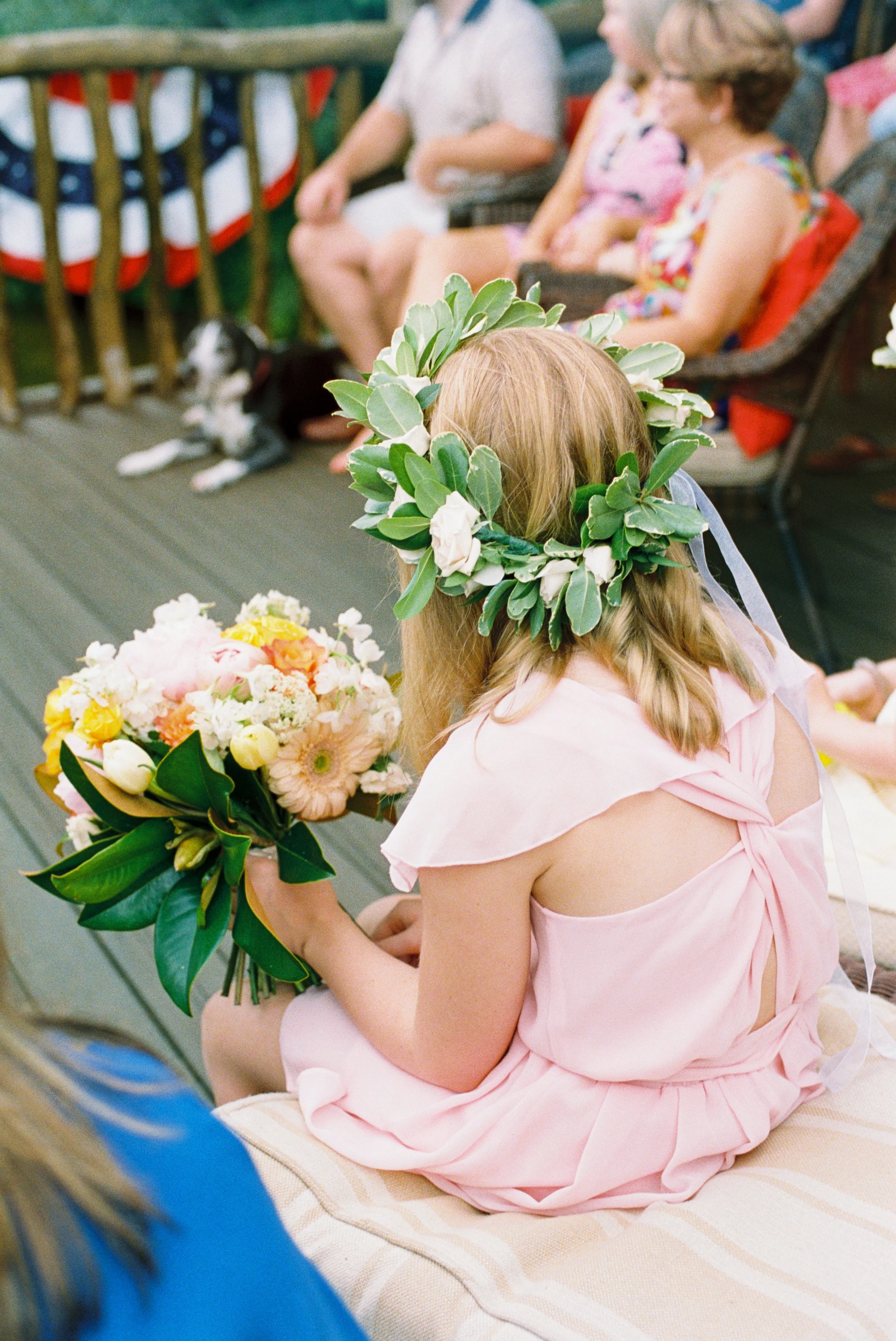 Flower girl in a floral crown holding a bouquet of yellow and white flowers, seated among wedding guests