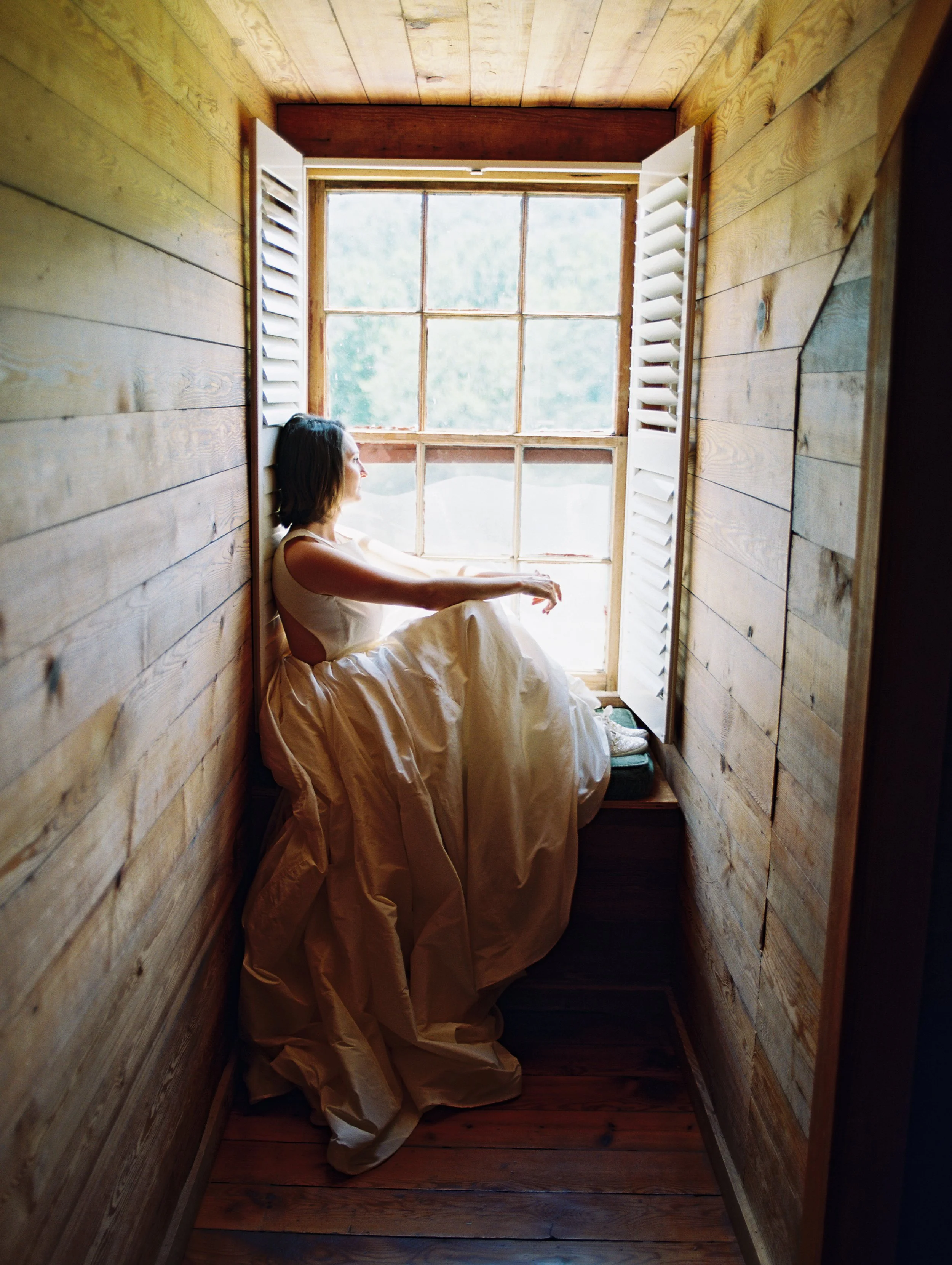 A woman sitting in a small wooden-walled nook by a window, wearing an ivory wedding dress, with legs pulled up and eyes closed, enjoying serenity, sunlight and a scenic view outside at Glen Ella Springs Hotel in Clarkesville, Georgia.