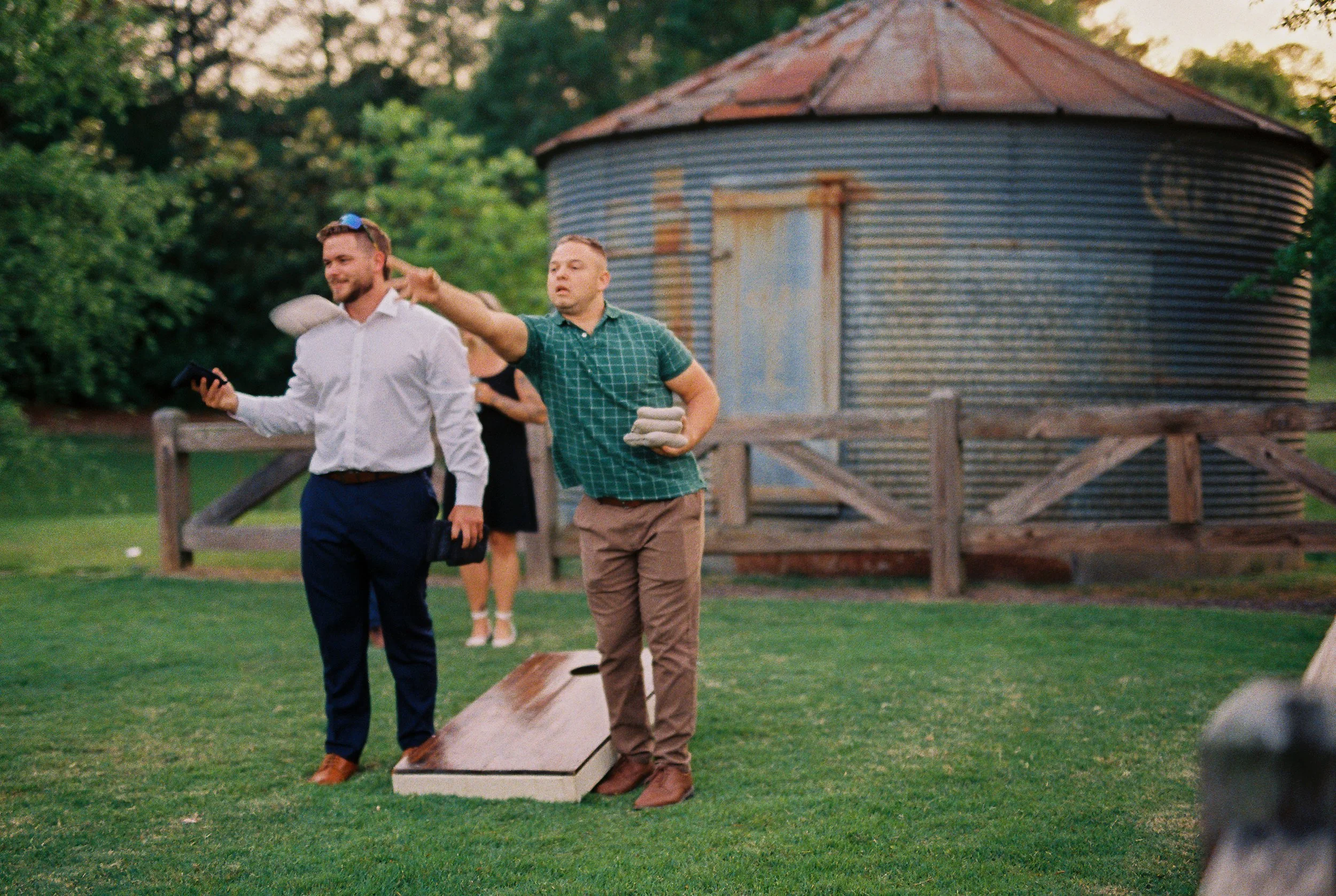 Male wedding guests playing cornhole on the lawn in front of a rustic corrugated metal silo