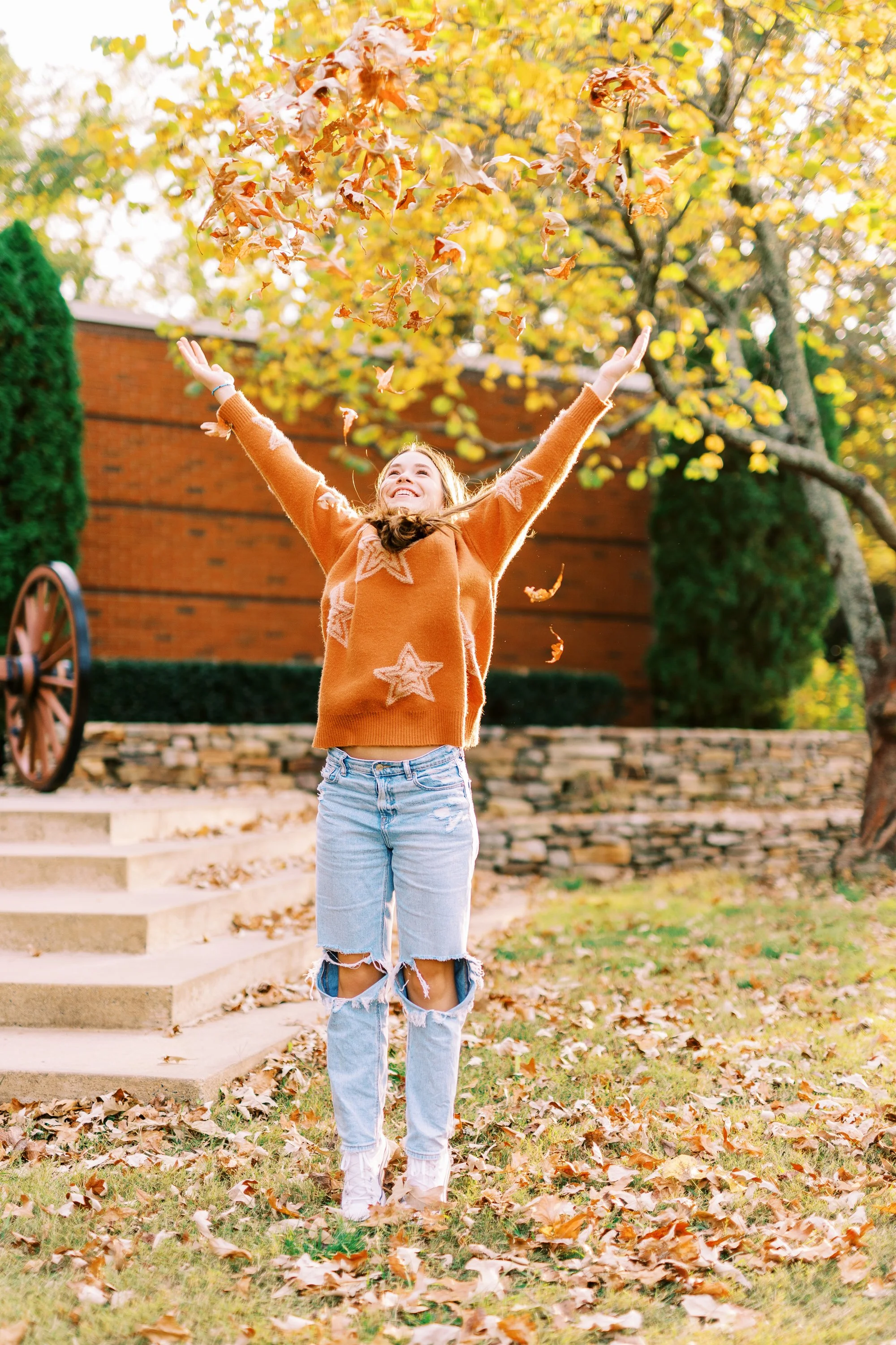 Senior portrait of Hannah with arms raised as leaves fall around her in an orange sweater at Kennesaw Mountain Battlefield Park in Kennesaw, Georgia