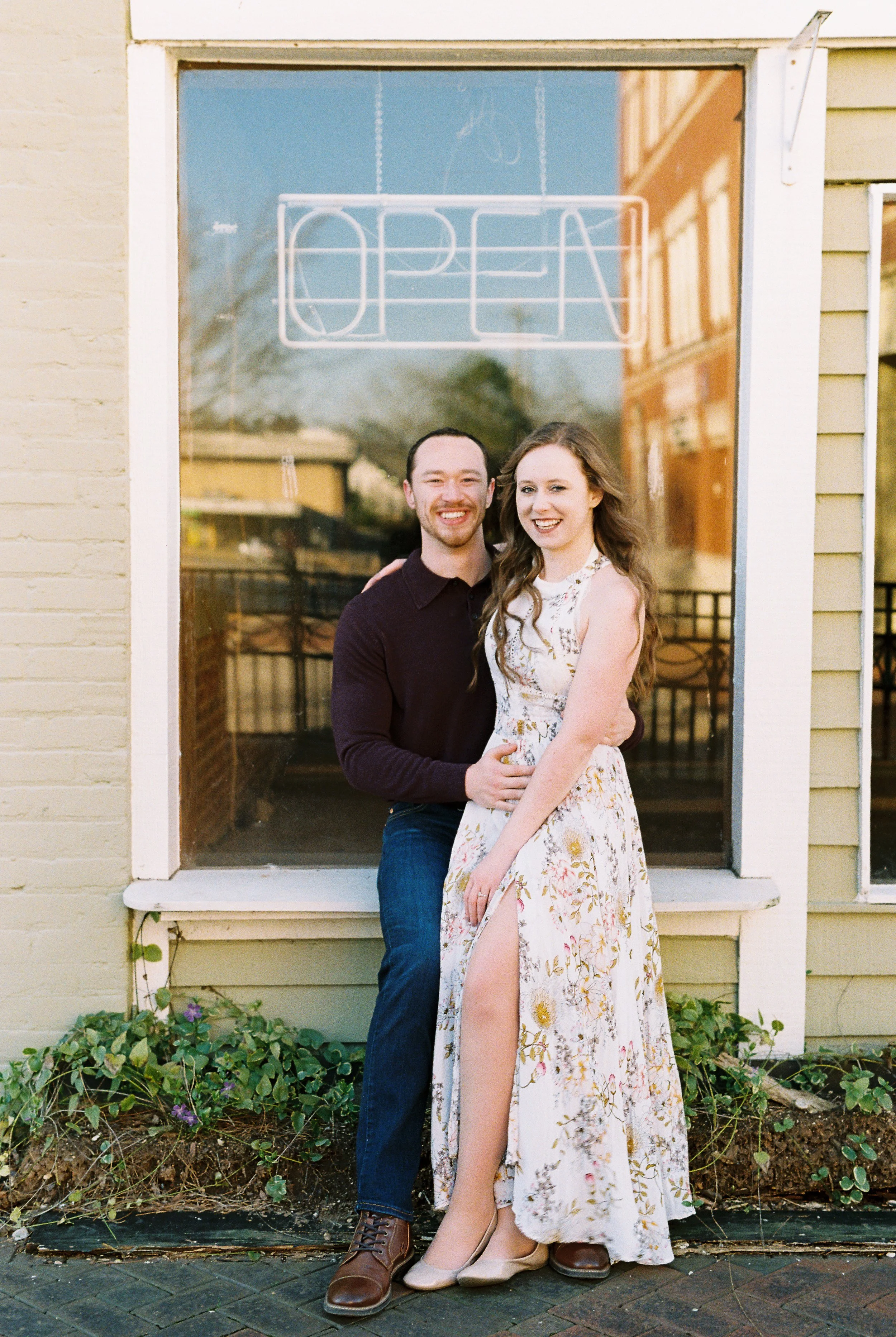 Conner and Maddie pose together in front of a shop window with a neon "Open" sign in downtown Dallas, Georgia.