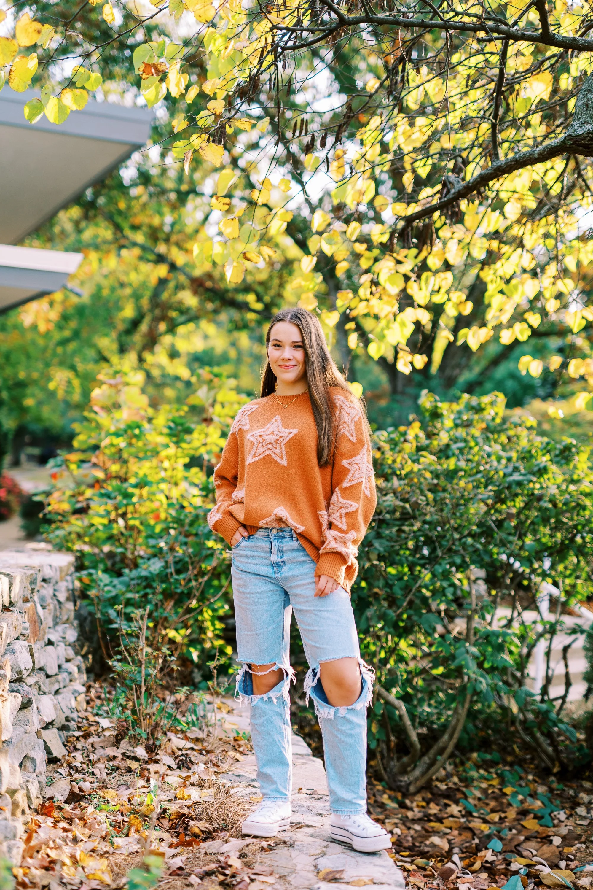 Senior portrait of Hannah standing beneath sunlit fall foliage in an orange sweater at Kennesaw Mountain Battlefield Park in Kennesaw, Georgia