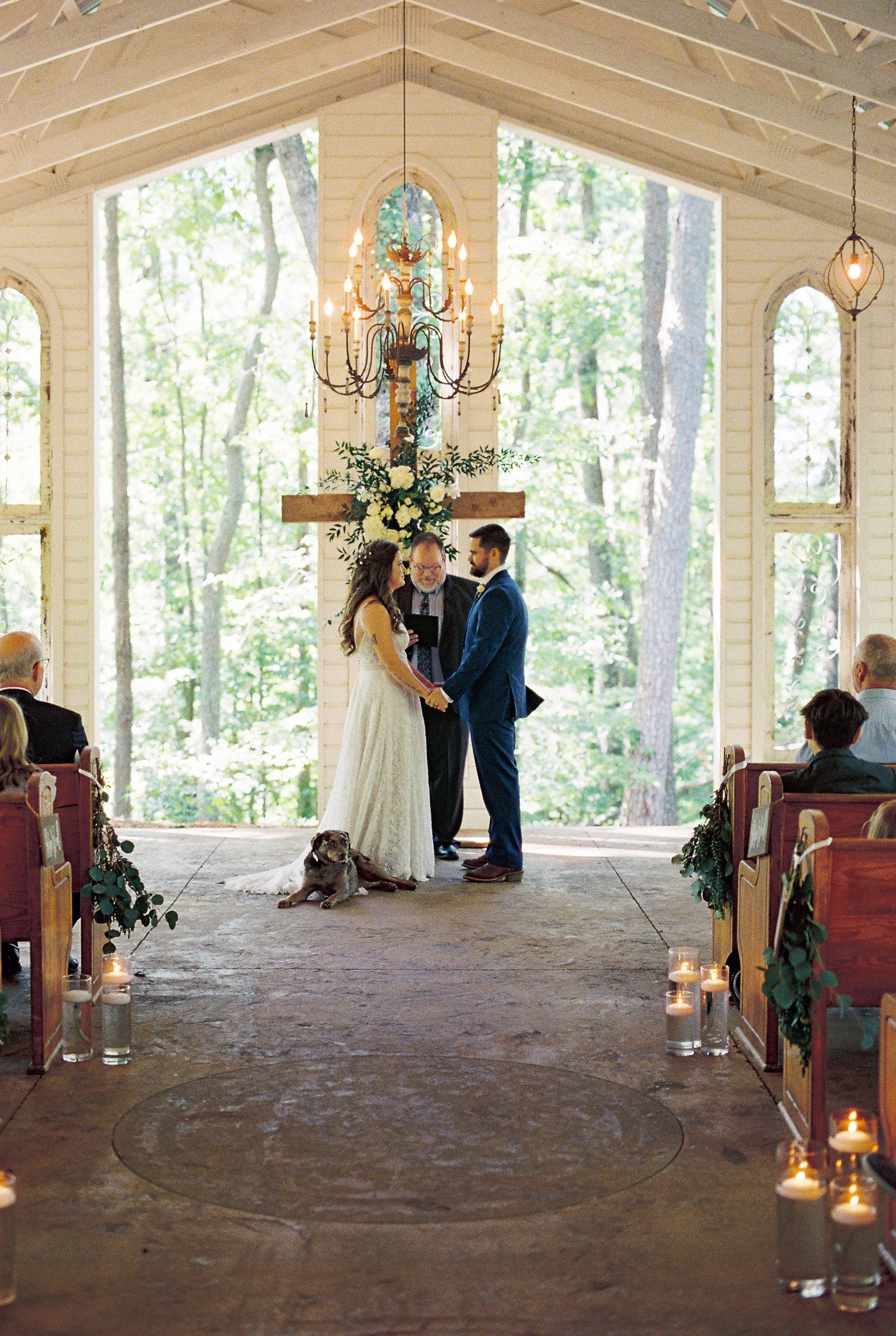Symmetrical view down the candlelit aisle toward the bride and groom exchanging vows at the altar beneath a wooden cross and chandelier, guests seated on both sides and Dakota resting near the altar