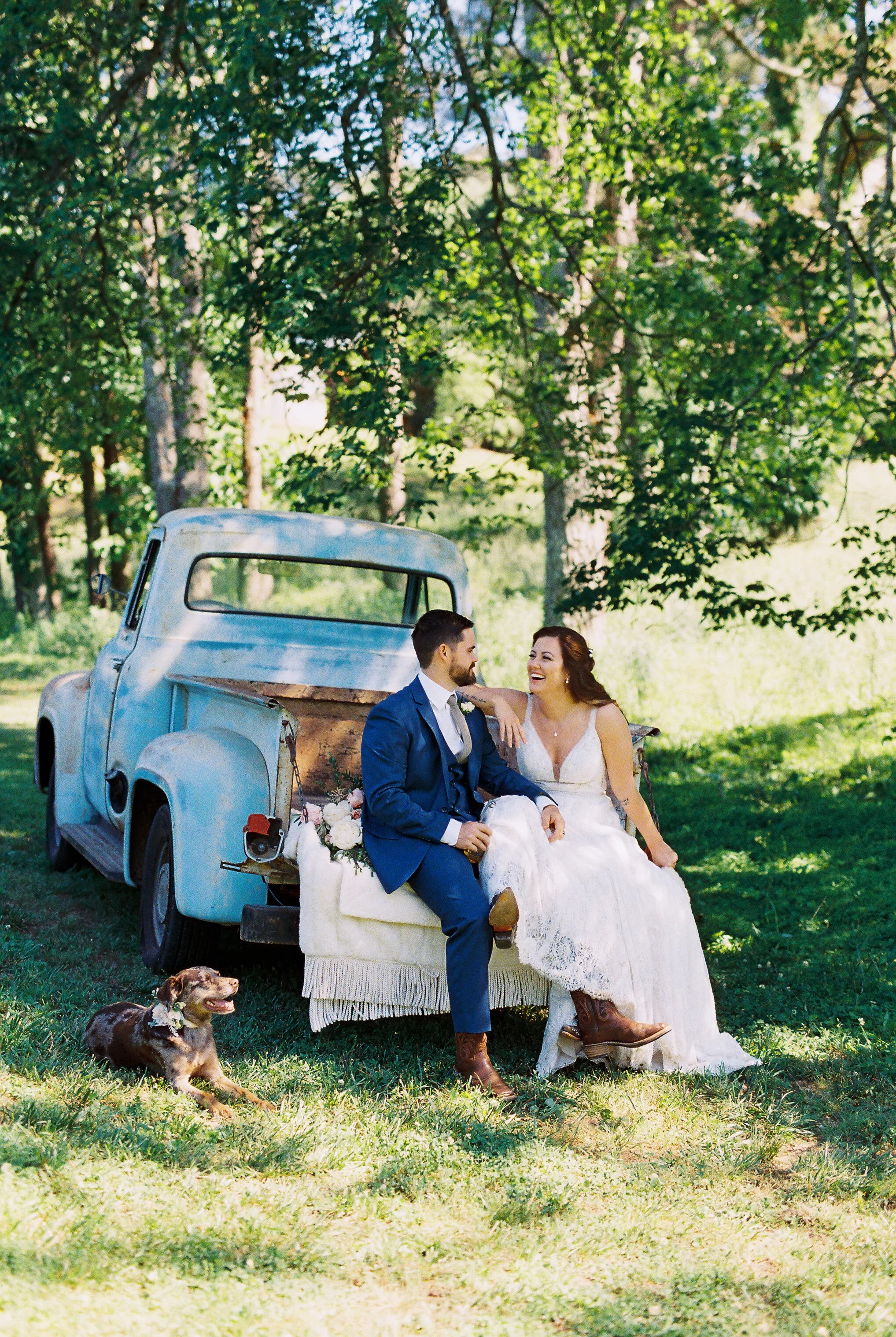 Bride and groom smiling at each other while seated on the vintage truck's tailgate, full truck in view with Dakota the dog lying at their feet