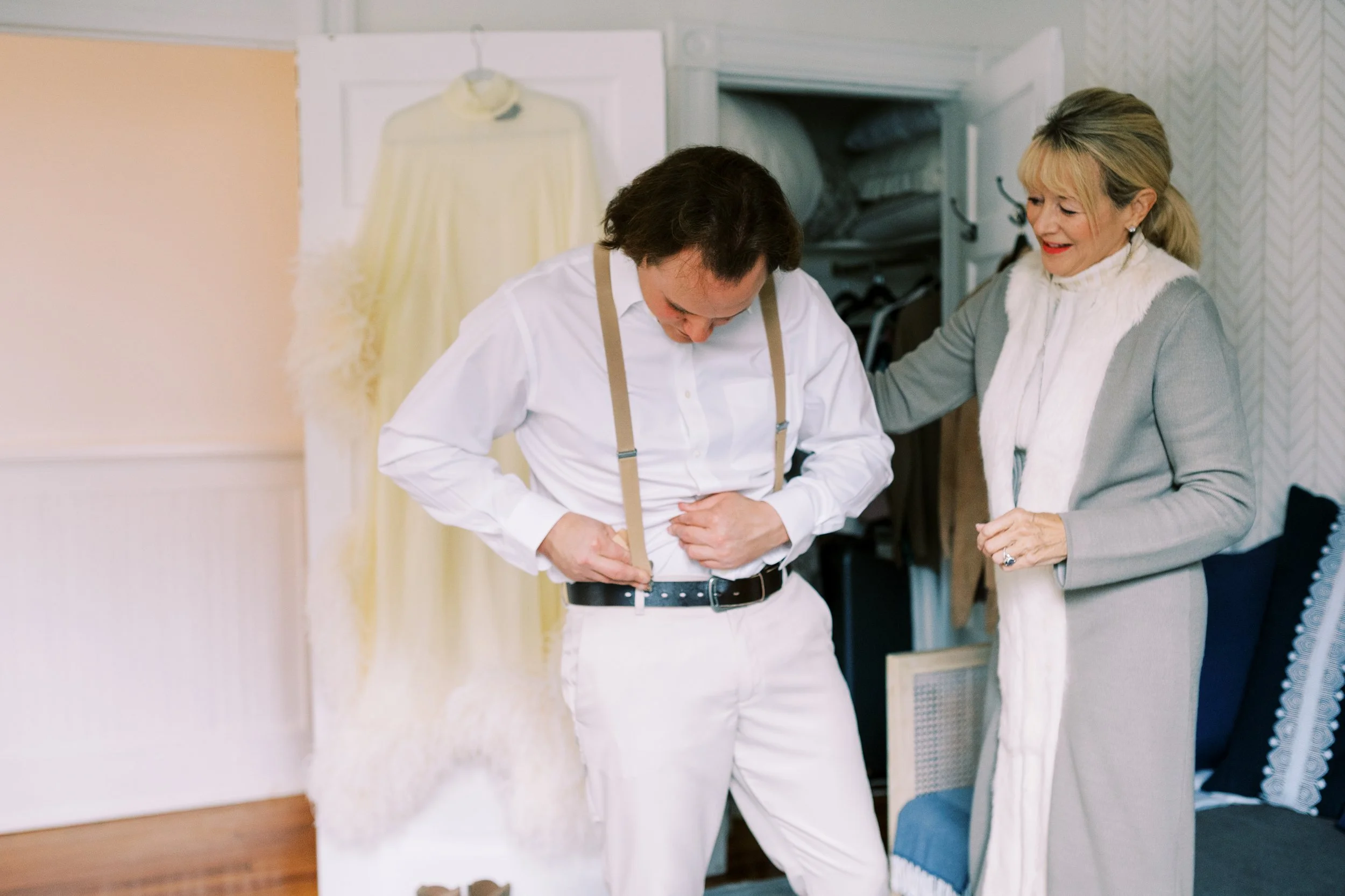 Groom putting on suspenders while getting ready with wedding dress hanging in the background