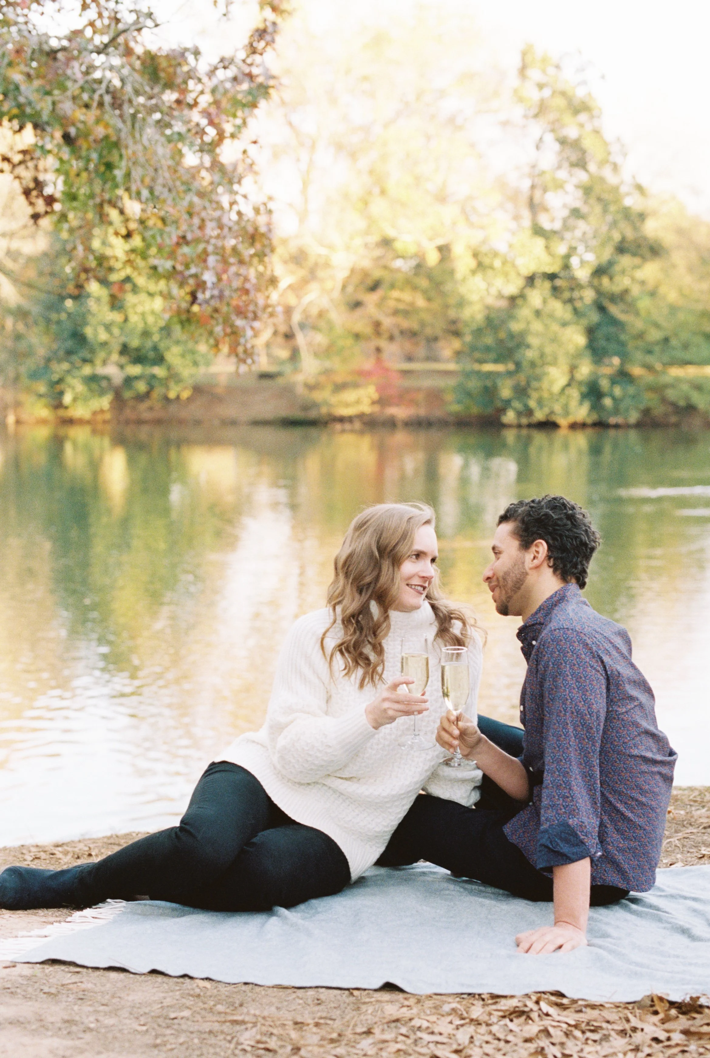 Couple seated on a picnic blanket by the lake, toasting champagne amid autumn foliage
