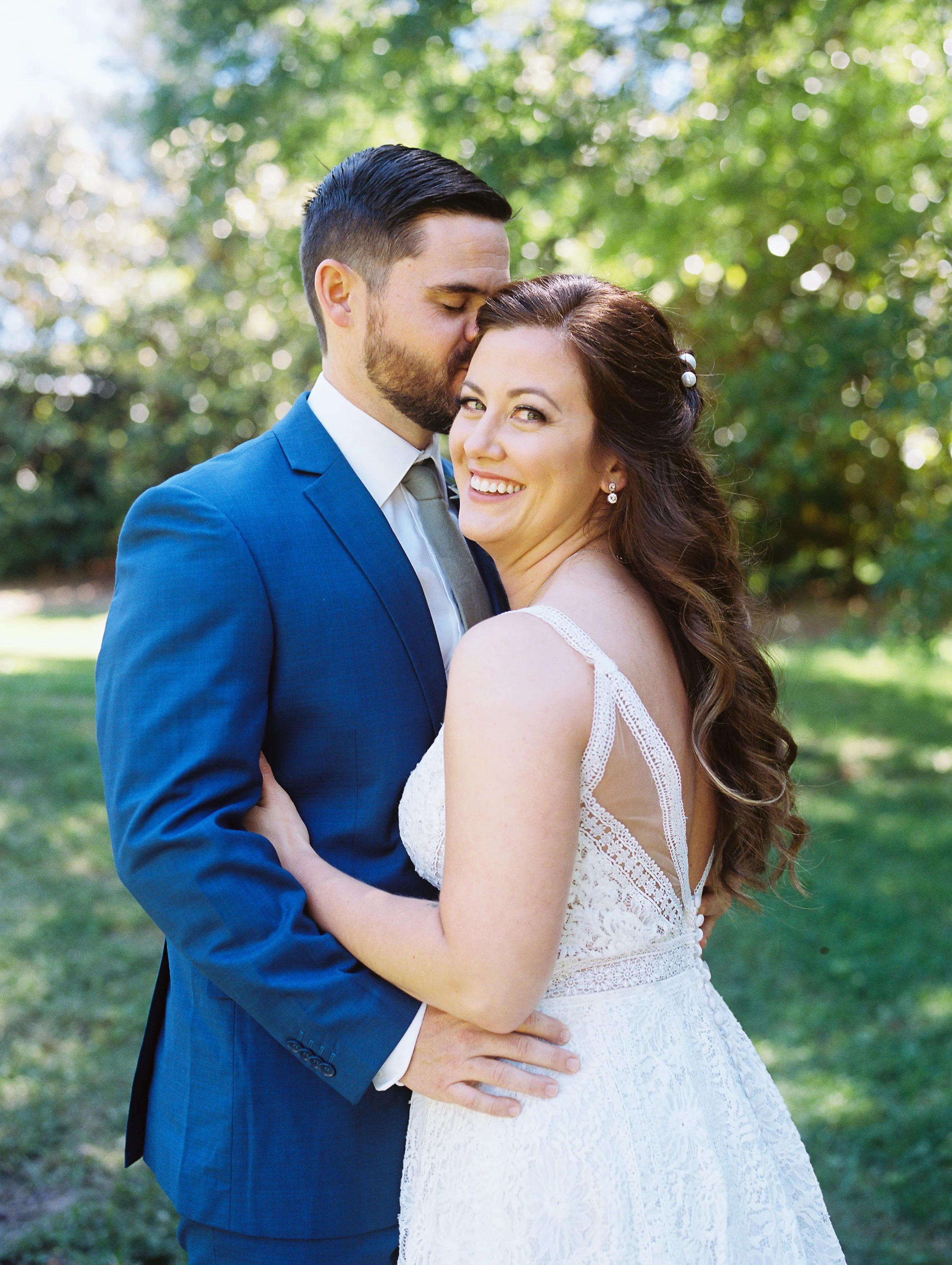 Groom kissing bride's forehead outdoors while she smiles at the camera