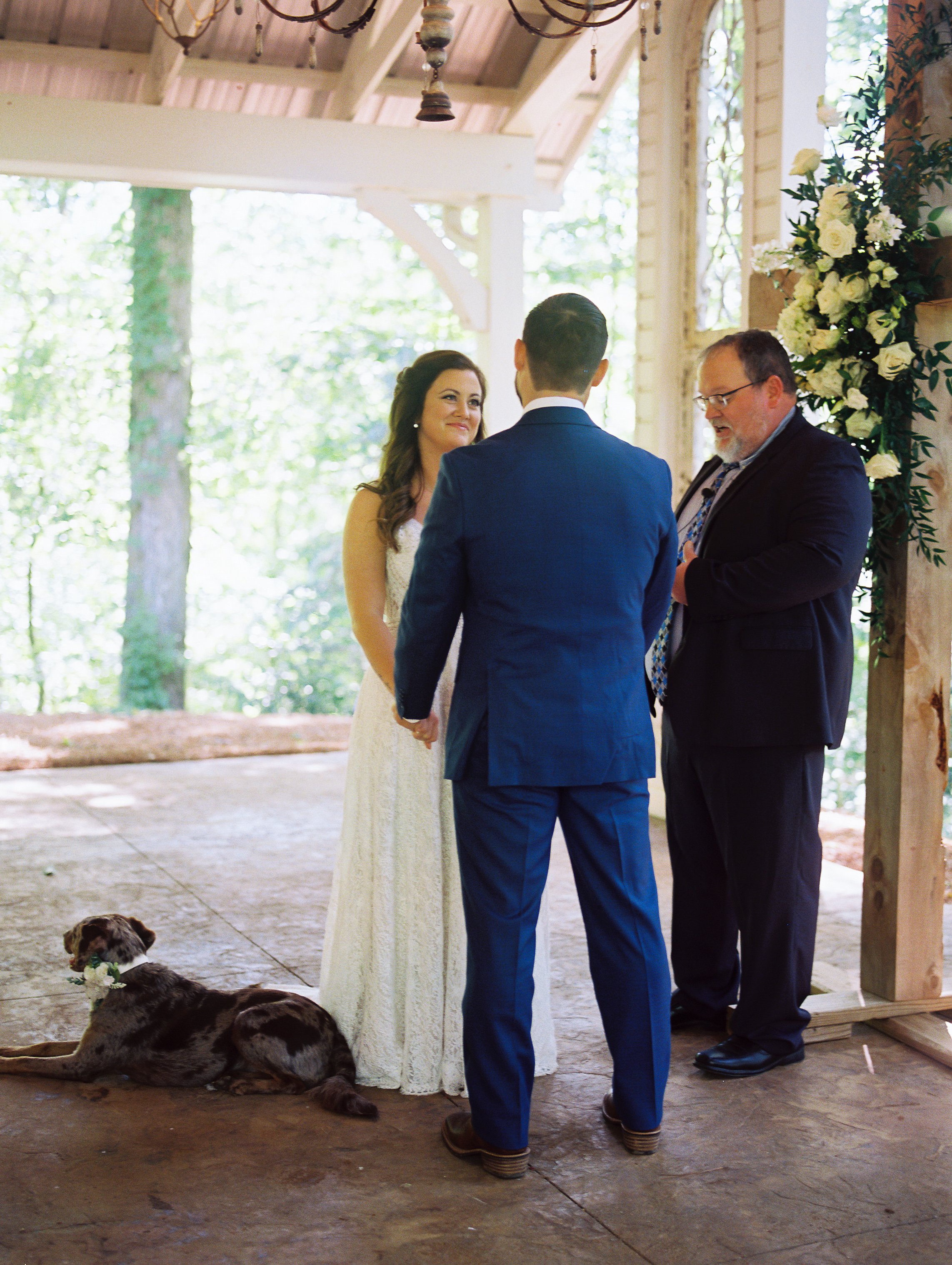 Bride and groom facing each other and holding hands at the altar during the ceremony, officiant standing beside them, Dakota resting on the ground nearby