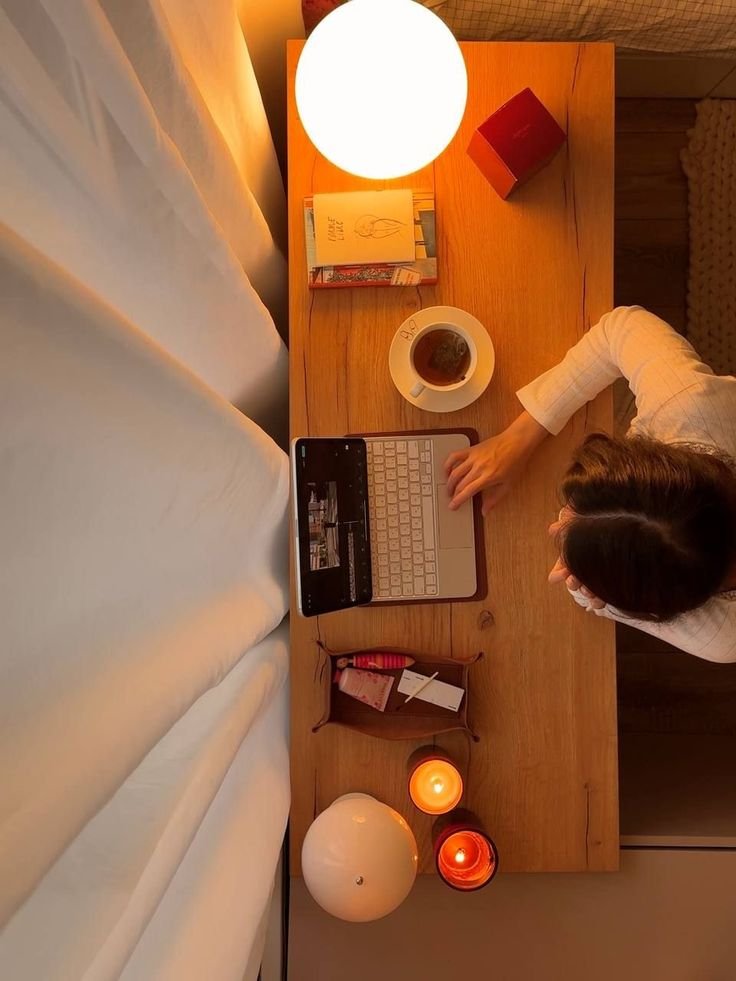 Overhead view of a person's workspace on a wooden desk with a glowing lamp, a cup of tea, a book, some candles, and a laptop. The person is typing on the laptop with their left hand.