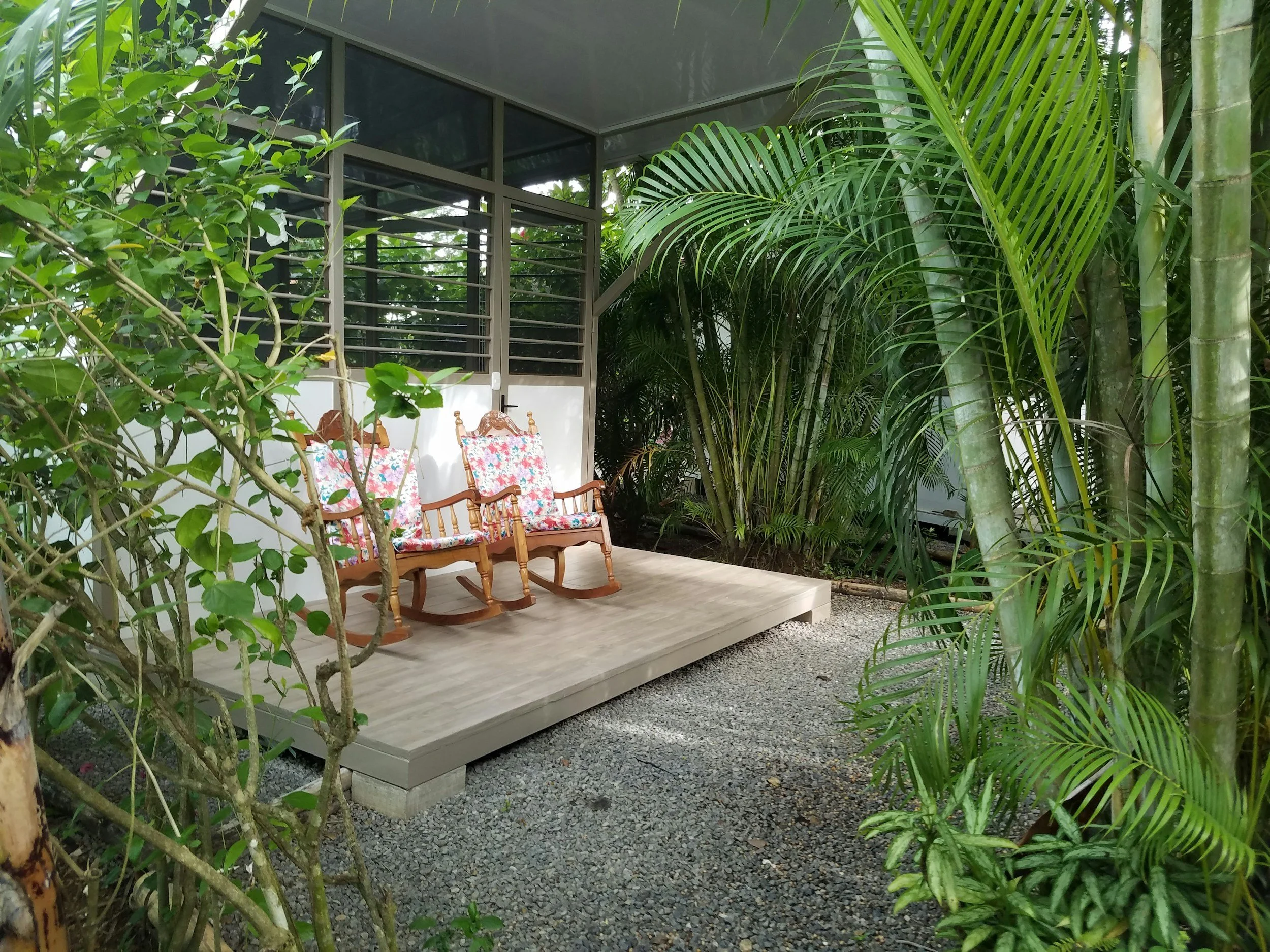 A wooden porch with two rocking chairs that have floral cushions, surrounded by lush green tropical plants and trees.