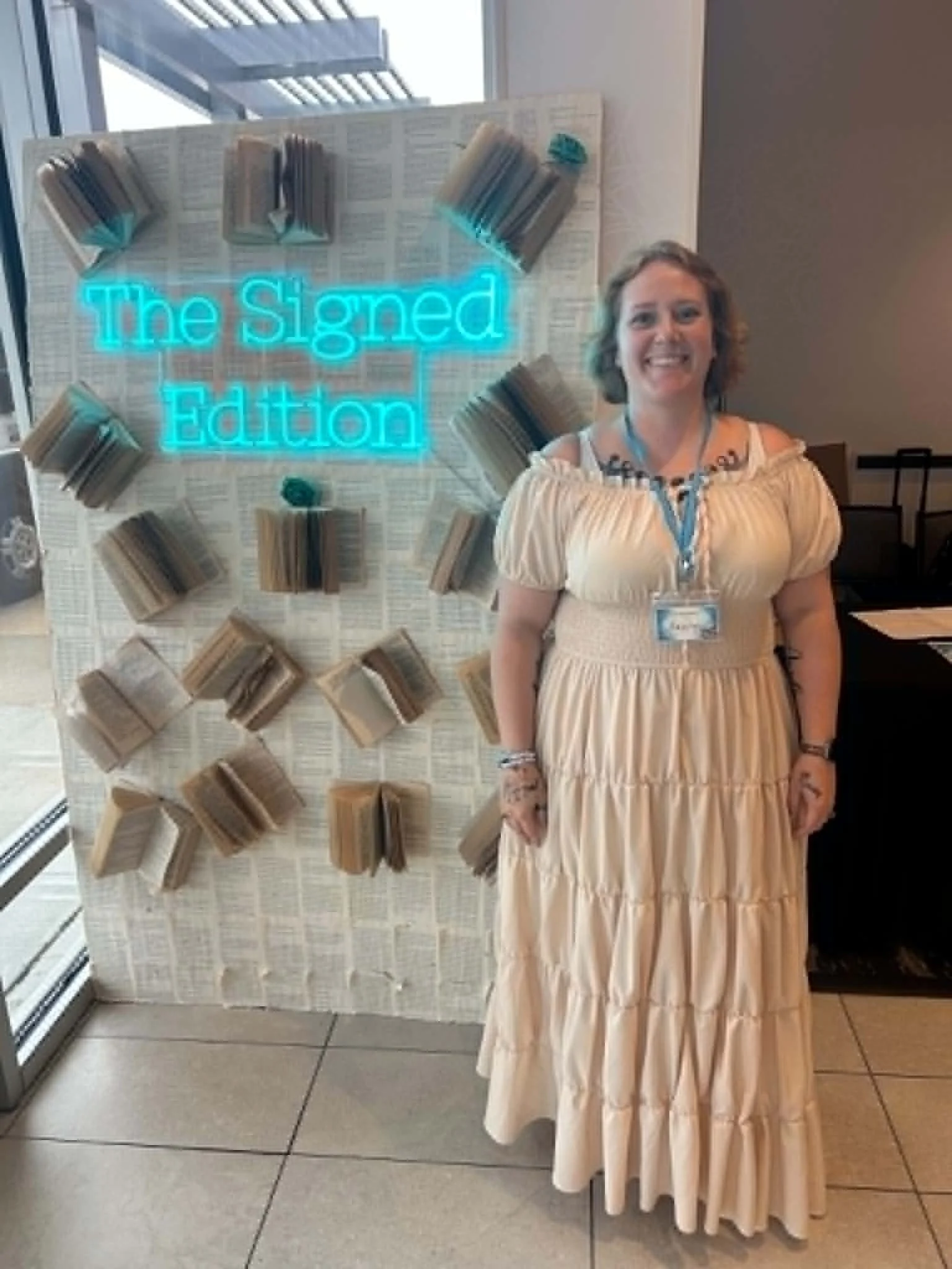A woman standing in front of a display with the neon sign 'The Signed Edition' and shelves with books. She is smiling, wearing a light-colored dress, and has a lanyard with an ID badge around her neck.