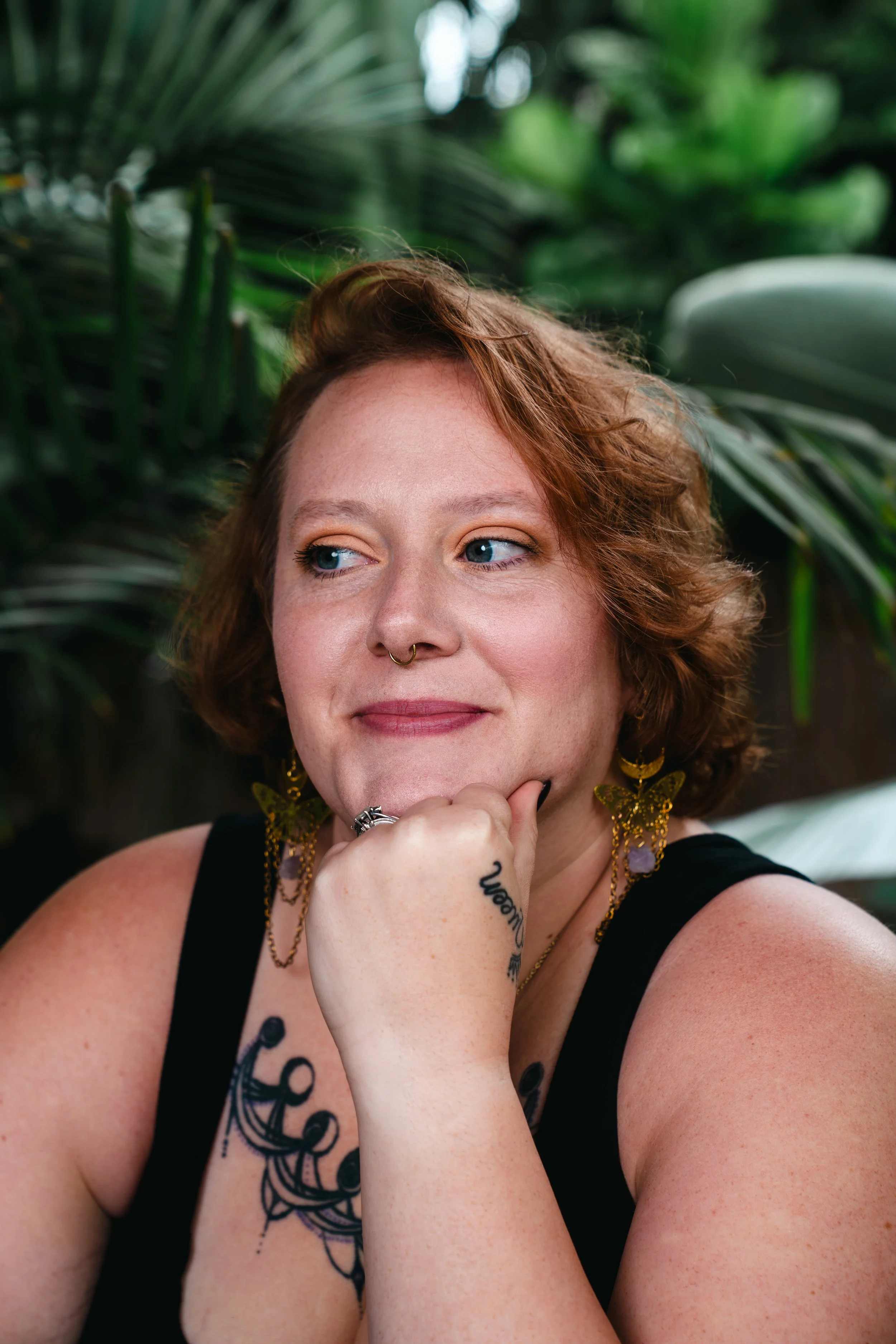 A woman with light skin, curly red hair, and blue eyes is resting her chin on her hand, wearing black clothing and jewelry, with palm trees in the background.