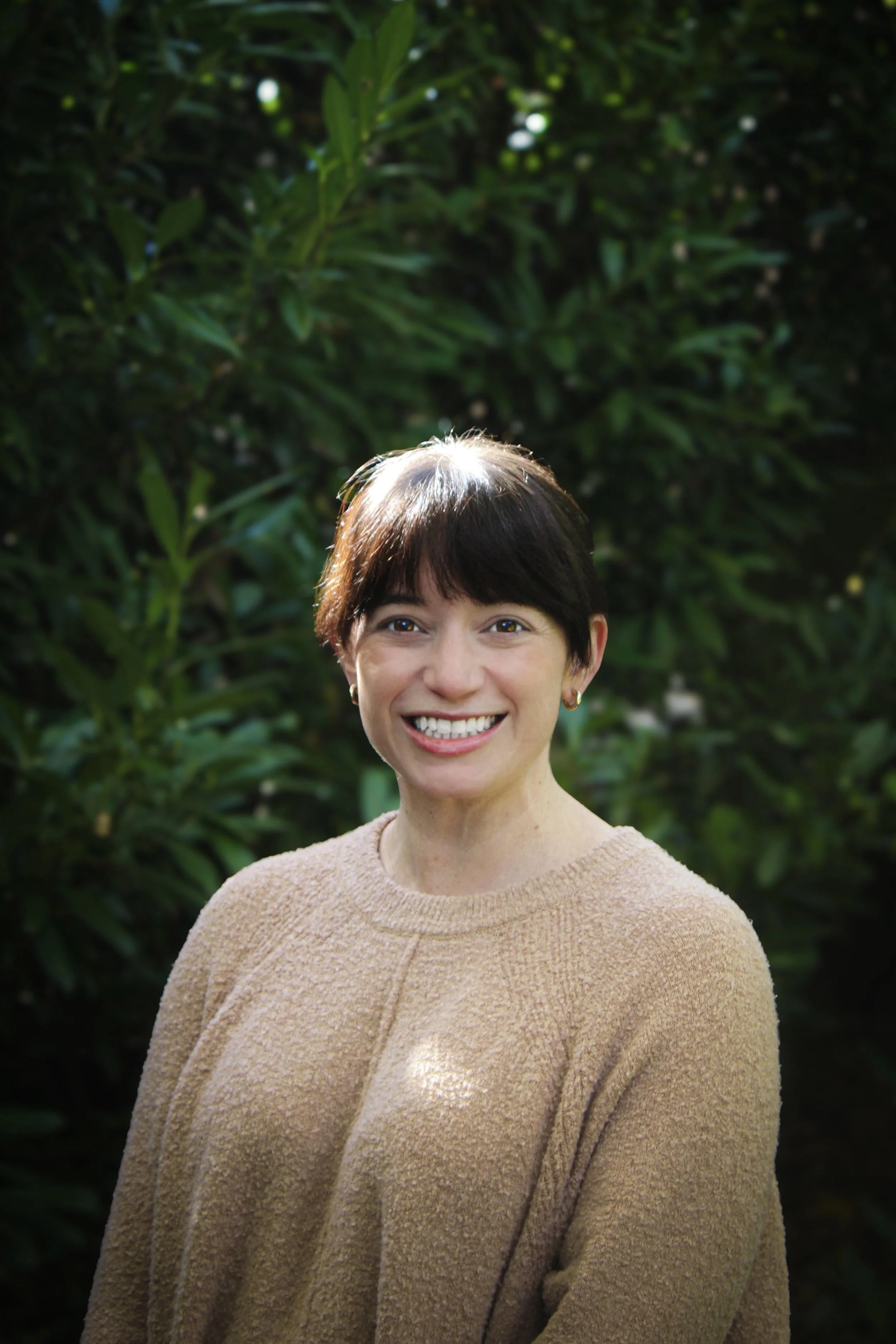 A woman with short brown hair and a beige sweater smiling outdoors with green foliage in the background.