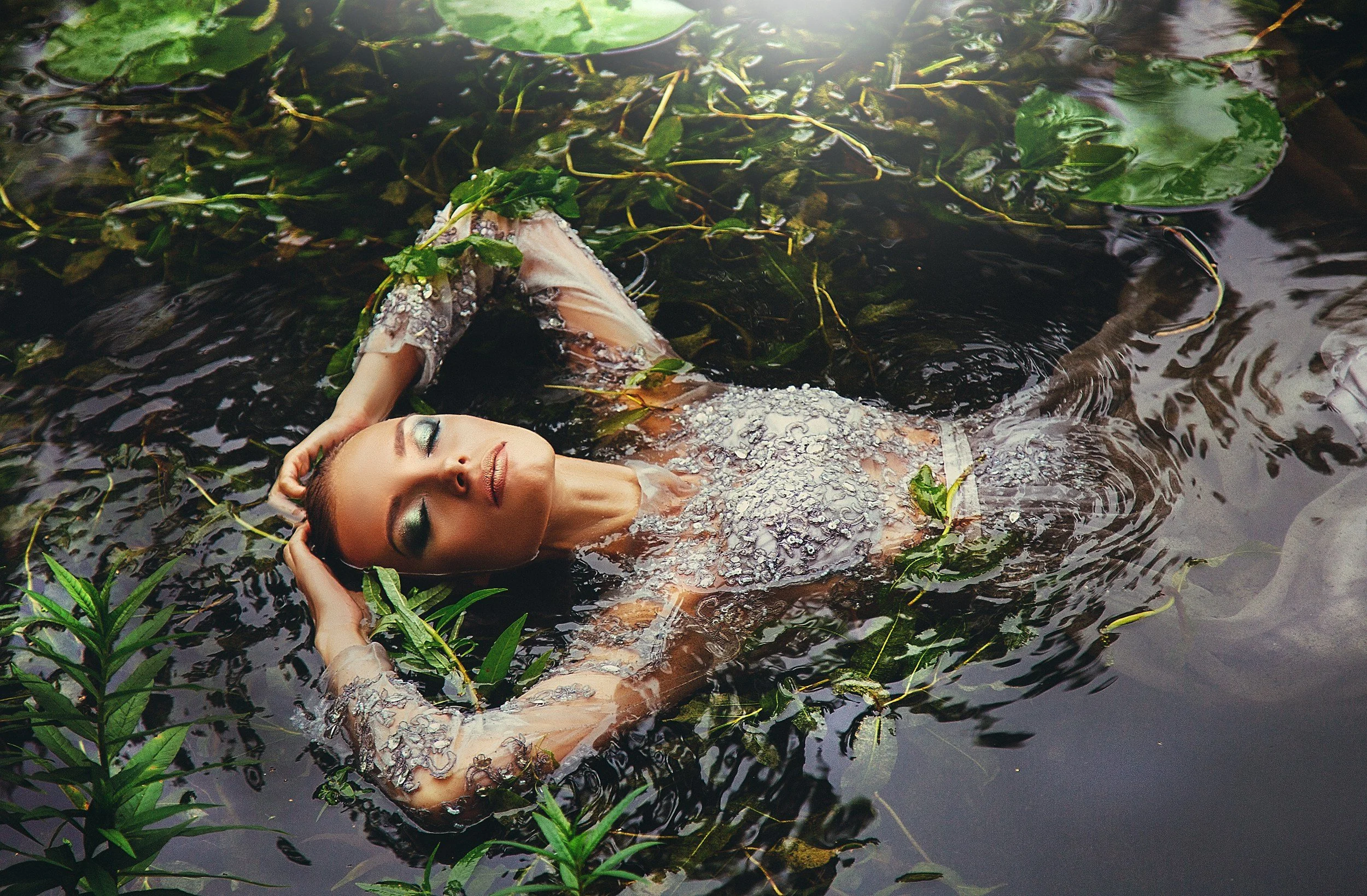 A woman with makeup and floral dress relaxing in water surrounded by green aquatic plants and lily pads, with her eyes closed and hands in her hair.