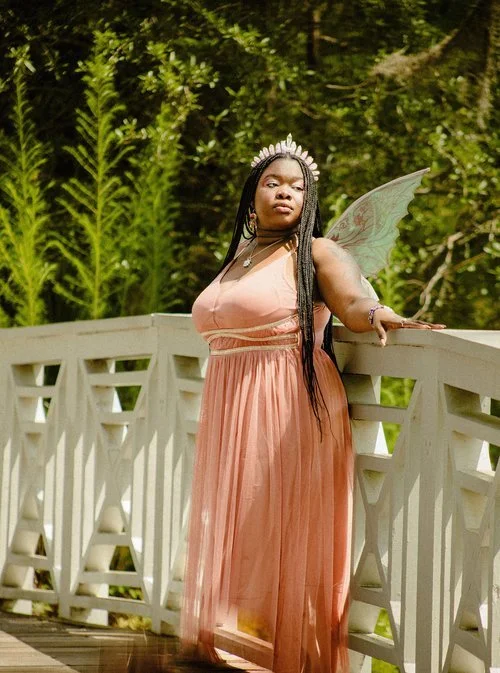 A woman dressed as a fairy with butterfly wings and a tiara, standing on a wooden bridge in a lush green forest.