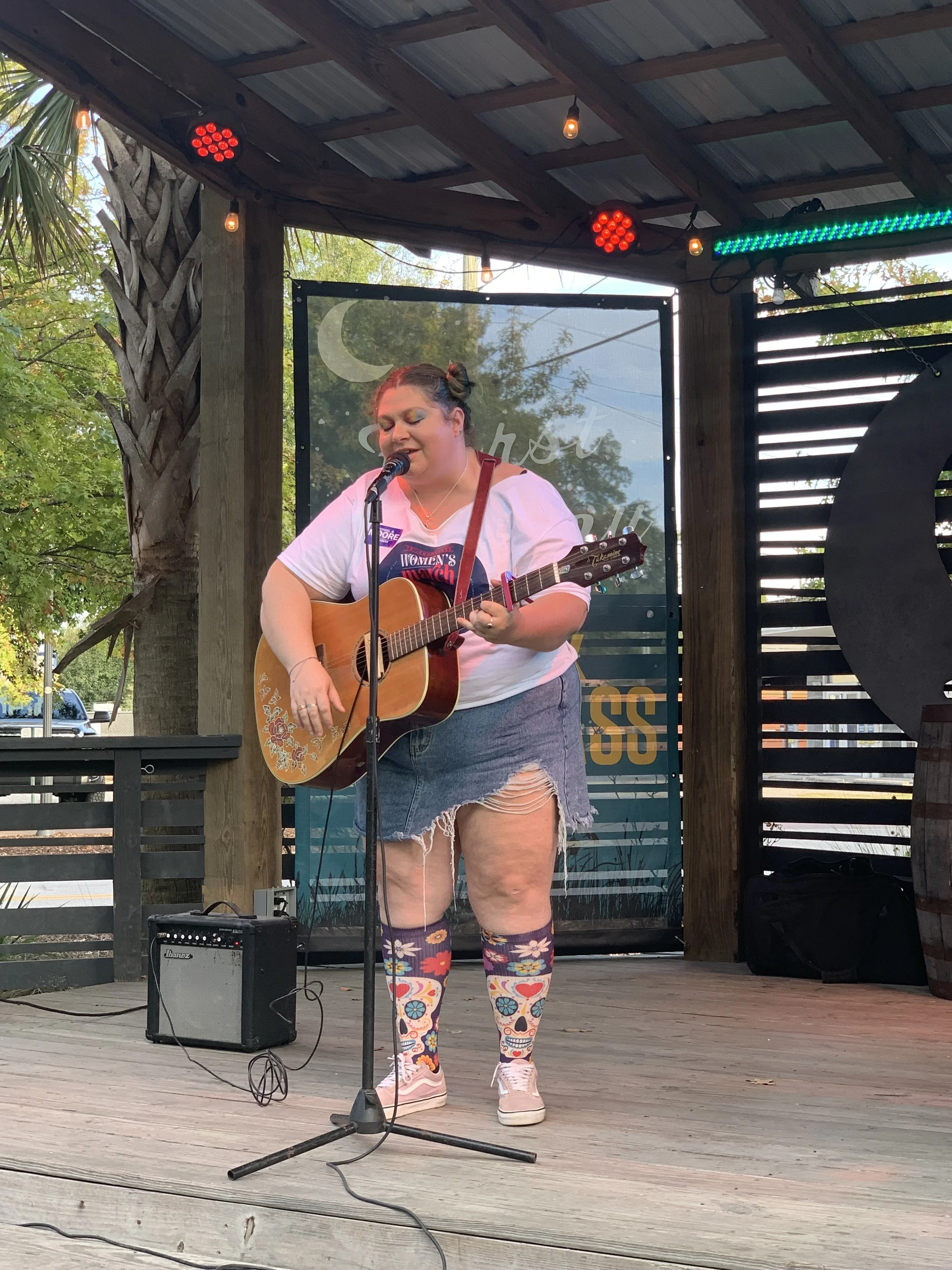A woman performing on stage with a guitar and singing into a microphone, wearing a white T-shirt, ripped denim shorts, and colorful, patterned knee-high socks, in an outdoor setting with string lights and a backdrop with moon and star designs.