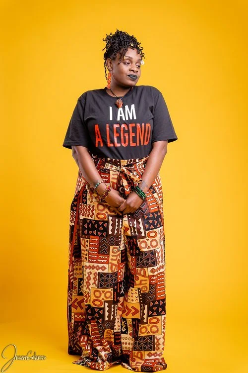A woman standing against a bright yellow background, wearing a black T-shirt that says 'I AM A LEGEND' in bold letters, and colorful patterned wide-leg pants. She has curly hair, large earrings, and is holding her hands in front of her.