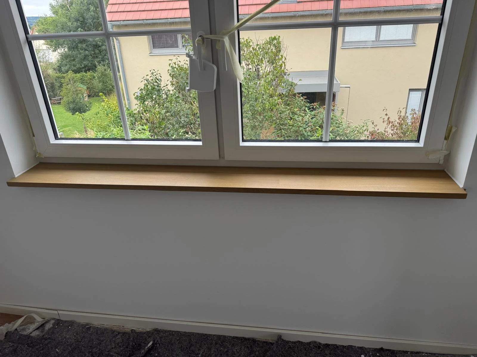 Ein Fenster mit einem braunen Fensterbrett, Blick nach draußen auf grüne Bäume und ein Nachbarhaus.