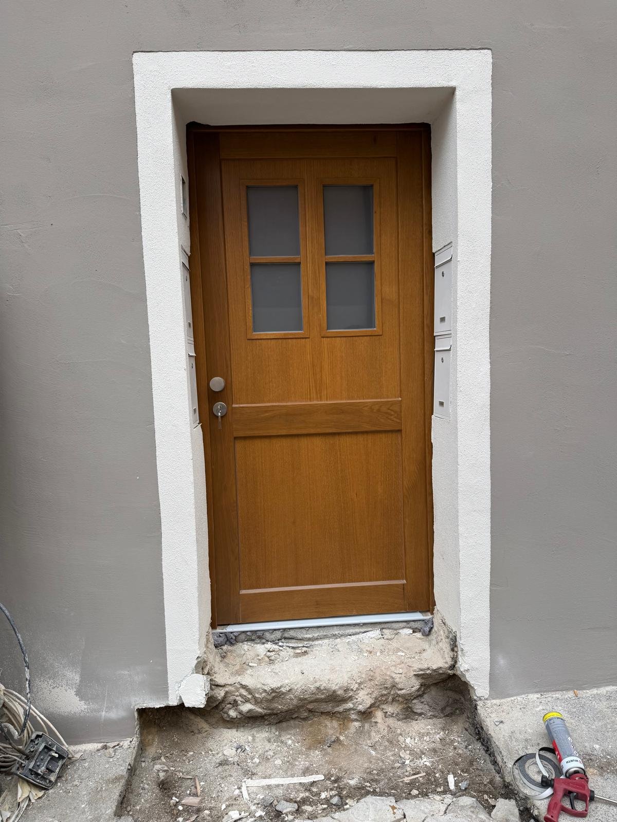 Ein Holztür mit vier Fenstern, eingearbeitet in eine weiße Türrahmen, im Bau befindliche Hausfassade mit Baustellenschutt und Werkzeug auf dem Boden.