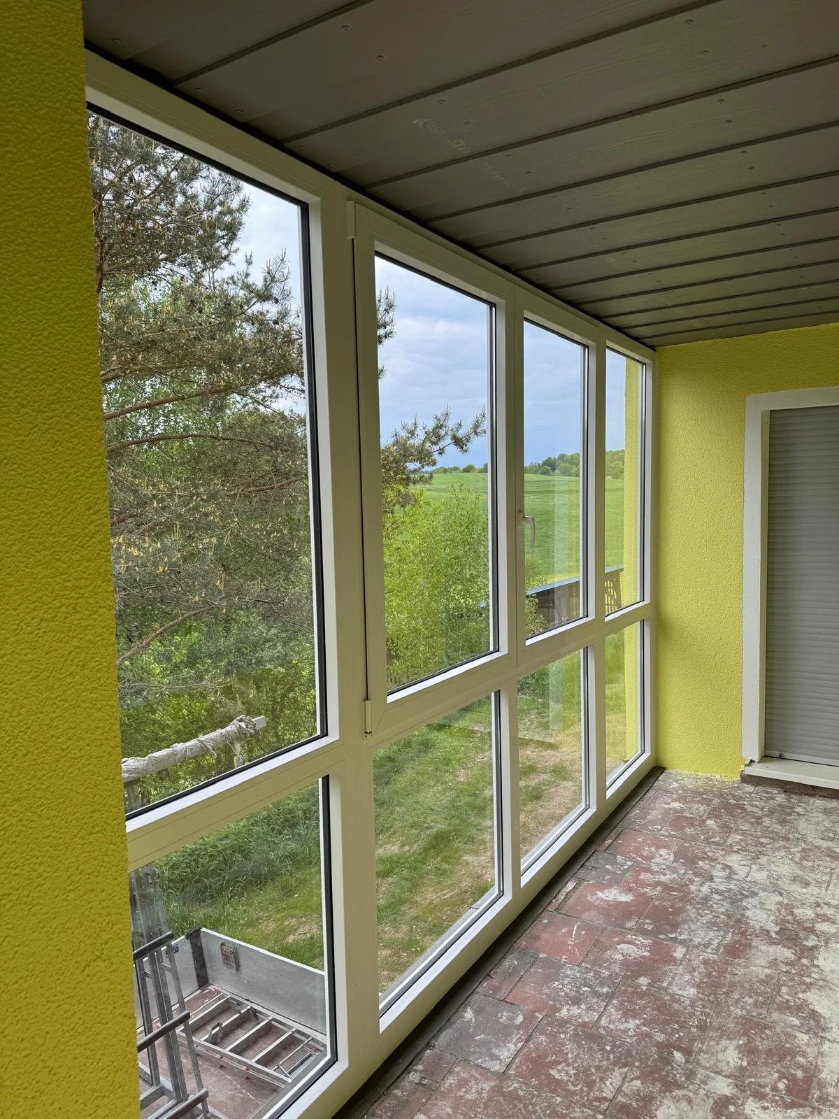 Innenansicht eines Balkons mit großen Fenstern, gelben Wänden, Fliesenboden und Blick auf grüne Landschaft.