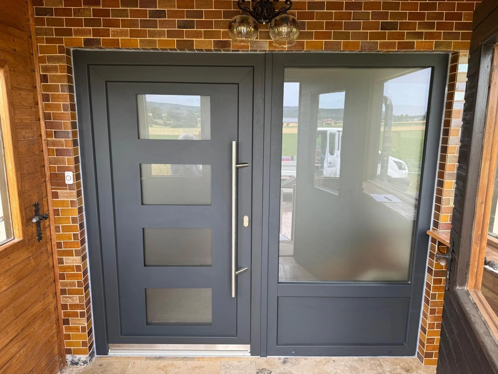 Moderne Eingangstür mit großen Glasfenstern, umgeben von Ziegel- und Holzverkleidung, mit Blick auf eine ländliche Landschaft.