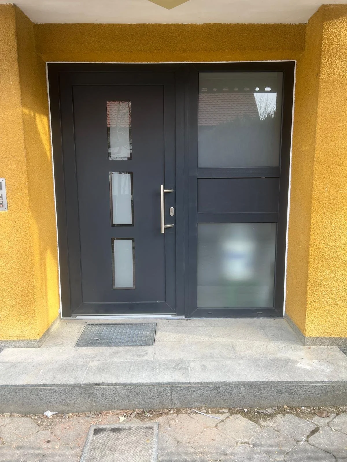 Eine schwarze Haustür mit drei kleinen quadratischen Glaseinsätzen und einem länglichen Türgriff, neben einem Fenster mit halb getöntem Glas, an einer gelben Wand. Vor der Tür liegt ein Pflasterstein.