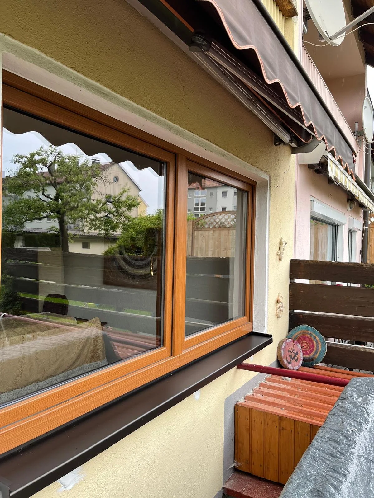 Balkon mit braunem Fensterrahmen, umlaufender Fensterbank, grauem Rollladen, Holzmöbeln, dekorativen Steinen, einem Baum in der Reflexion und umliegenden Gebäuden.
