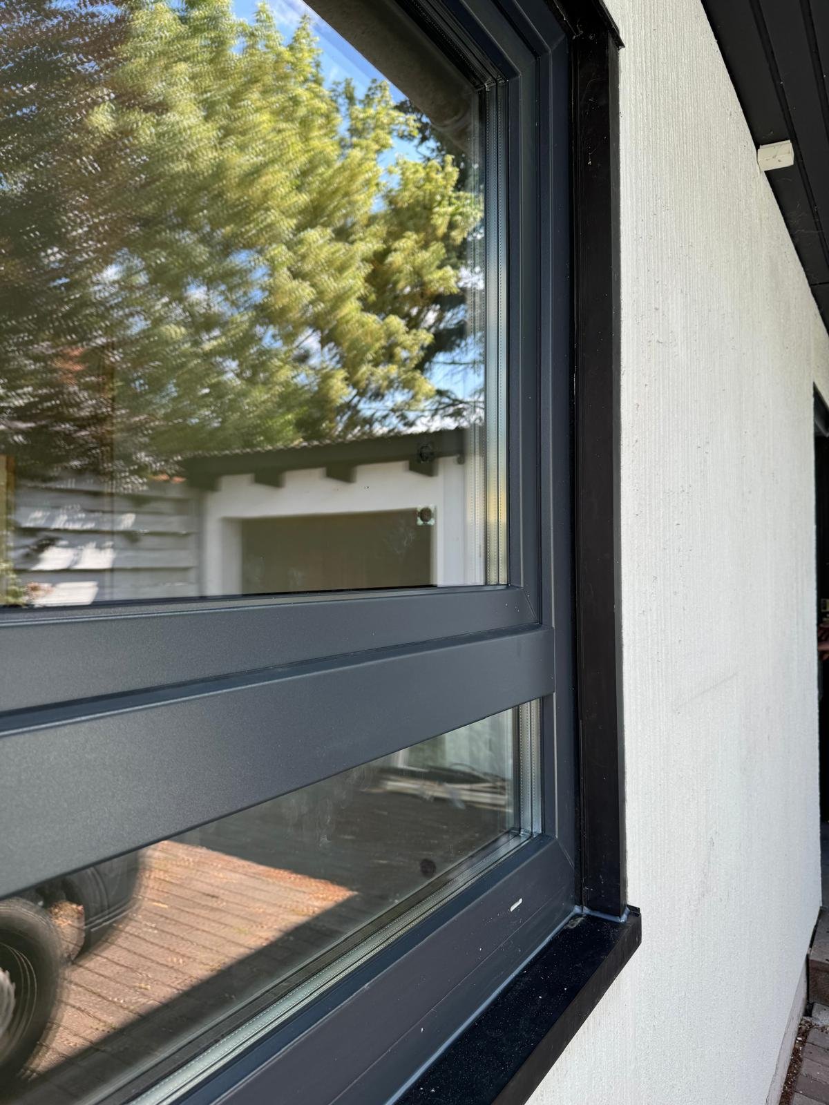 Ansicht eines modernen Fensters mit schwarzem Rahmen, eingebaut in eine weiße Hauswand, mit Blick auf einen Garten mit grünen Bäumen und eine Terrasse.