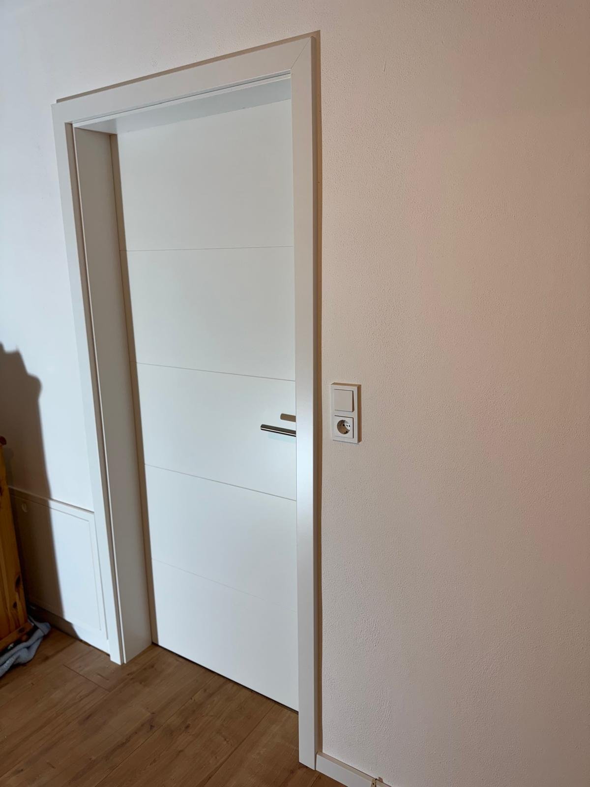 Weiße moderne Tür in einem Raum mit Holzboden, daneben eine weiße Wand mit Steckdose.