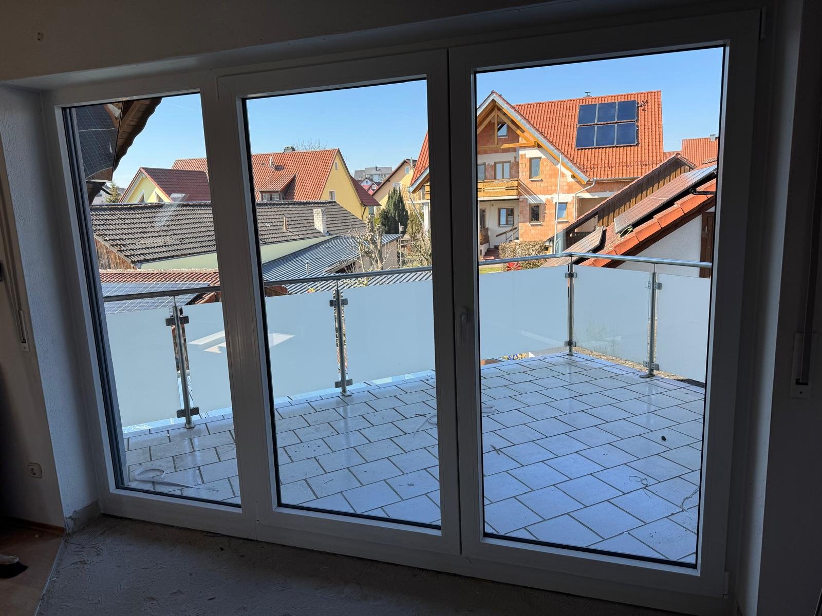 Blick von einem Innenraum durch eine große Balkon-Tür auf einen Balkon mit Pflastersteinen und eine Glasgeländer, sowie Nachbarhäuser mit roten und grauen Dächern und Solarzellen, bei blauem Himmel.