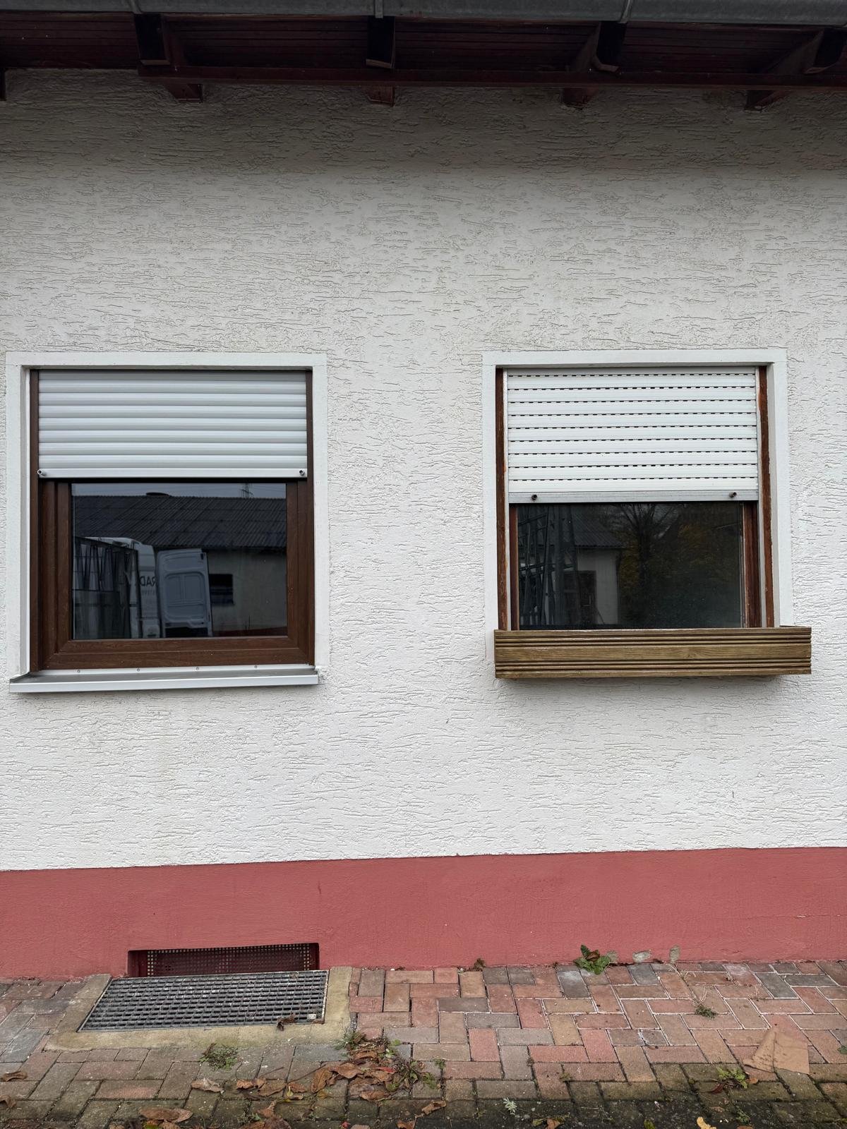 Zwei Fenster mit Rollläden an einer weißen Hauswand, eine mit Holzrahmen und eine mit einer Holzvase darunter. Vor dem Haus liegt ein gepflasterter Gehweg mit herabgefallenen Blättern.