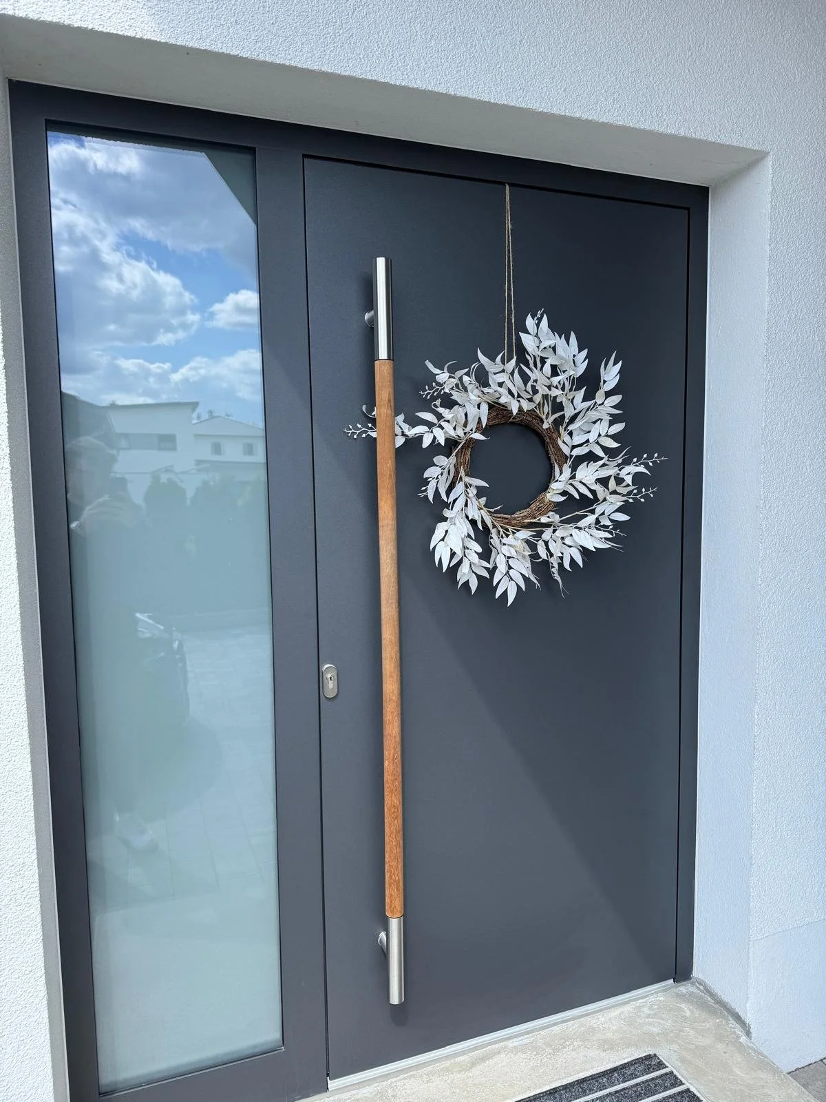 Moderne Haustür in Dunkelgrau mit silbernem langen Türgriff, verzierte Türkranz mit weißen Blättern, im Hintergrund gläserne Fenster, reflektierende Wolken am Himmel.