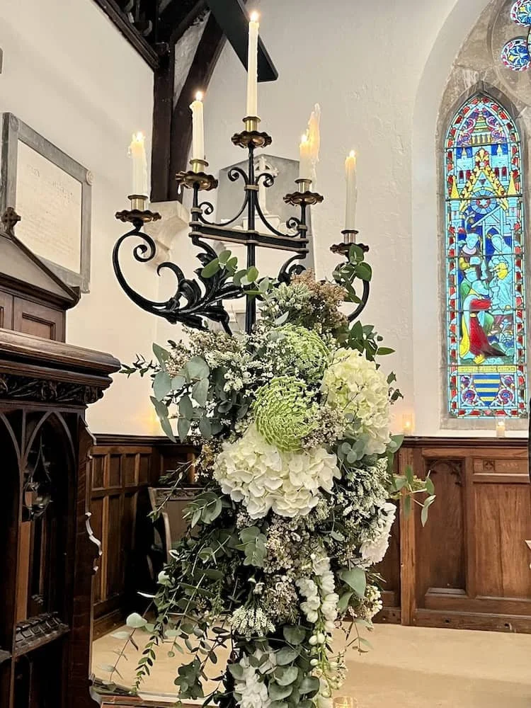 St Catherine's  Cultural Centre Kinsale interior with a black chandelier holding lit candles, decorated with a large floral arrangement of white hydrangeas, roses, and green foliage, and a stained glass window in the background.