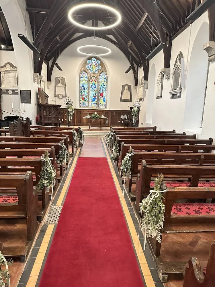 Interior of St Catherine's with wooden pews, floral aisle decorations, a red carpet, stained glass window, and circular ceiling lights.