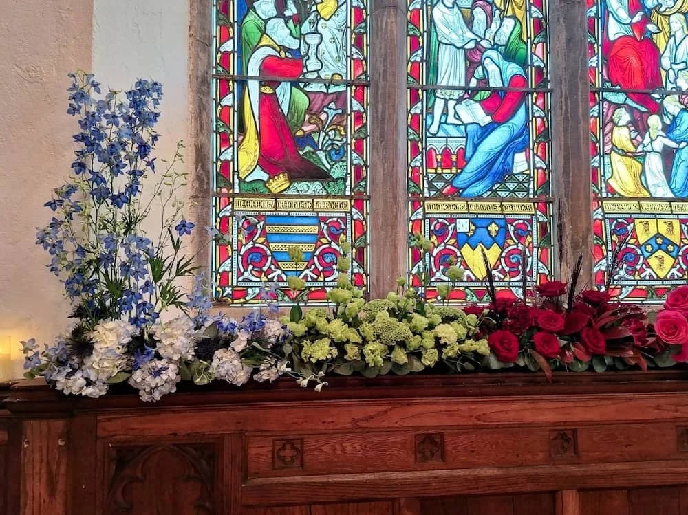 Colorful stained glass window in St Catherine's  Cultural Centre Kinsale with a floral arrangement on a wooden ledge below it, featuring blue, white, and red flowers.