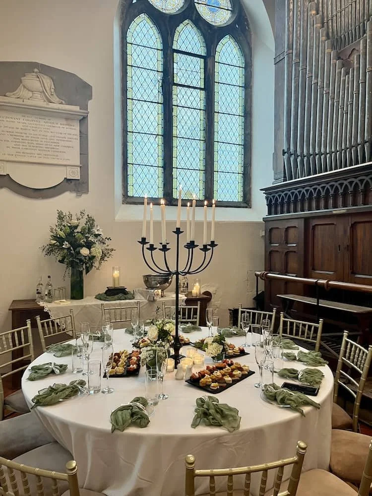 Round table set for a formal event with candles, floral arrangements, and appetisers, in St Catherine's  Cultural Centre Kinsale with stained glass windows and wood paneling.