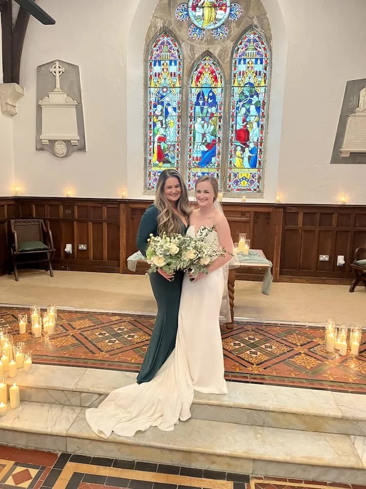 Two women in wedding attire standing in St Catherine's  Cultural Centre Kinsale with stained glass windows, candles, and wooden pews, holding a bouquet of flowers.