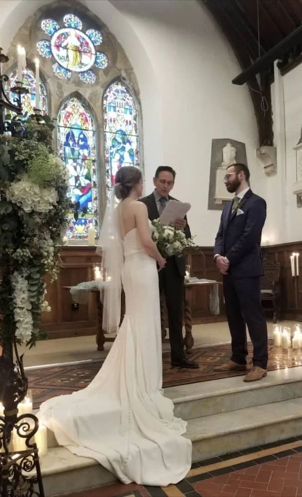 A bride and groom standing during their wedding ceremony in St Catherine's  Cultural Centre Kinsale, with an officiant reading from a paper. The bride is wearing a white wedding gown, and the groom is wearing a dark suit.