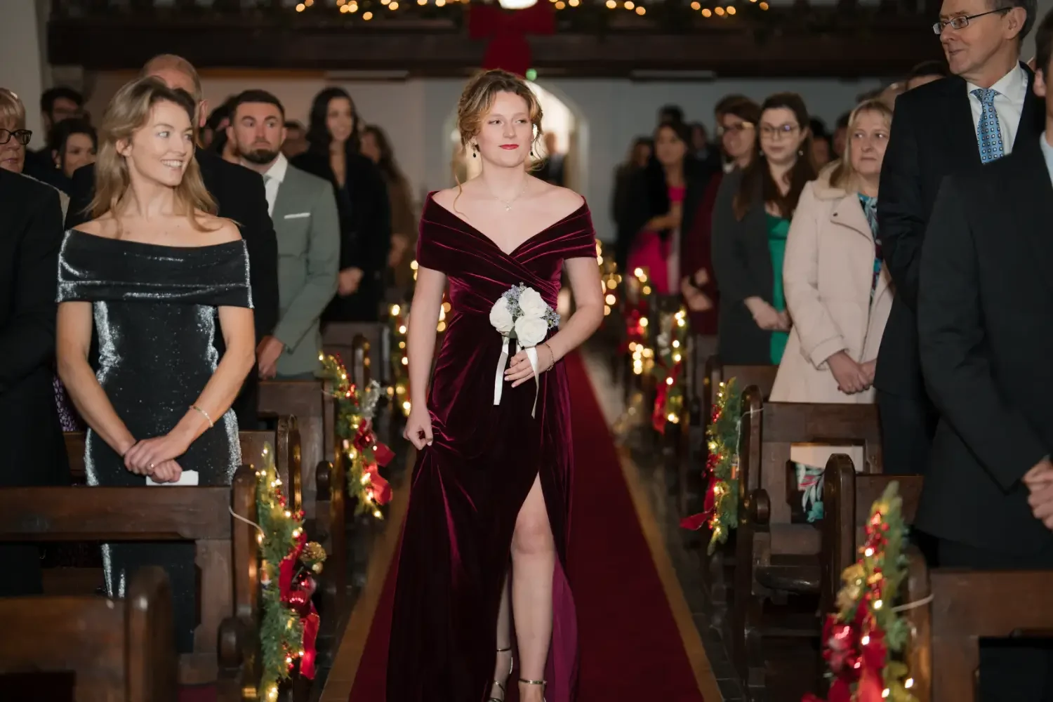 A Christmas Bride in a burgundy dress walks down a flower-lined aisle in St Catherine's, Kinsale, surrounded by seated and standing guests.