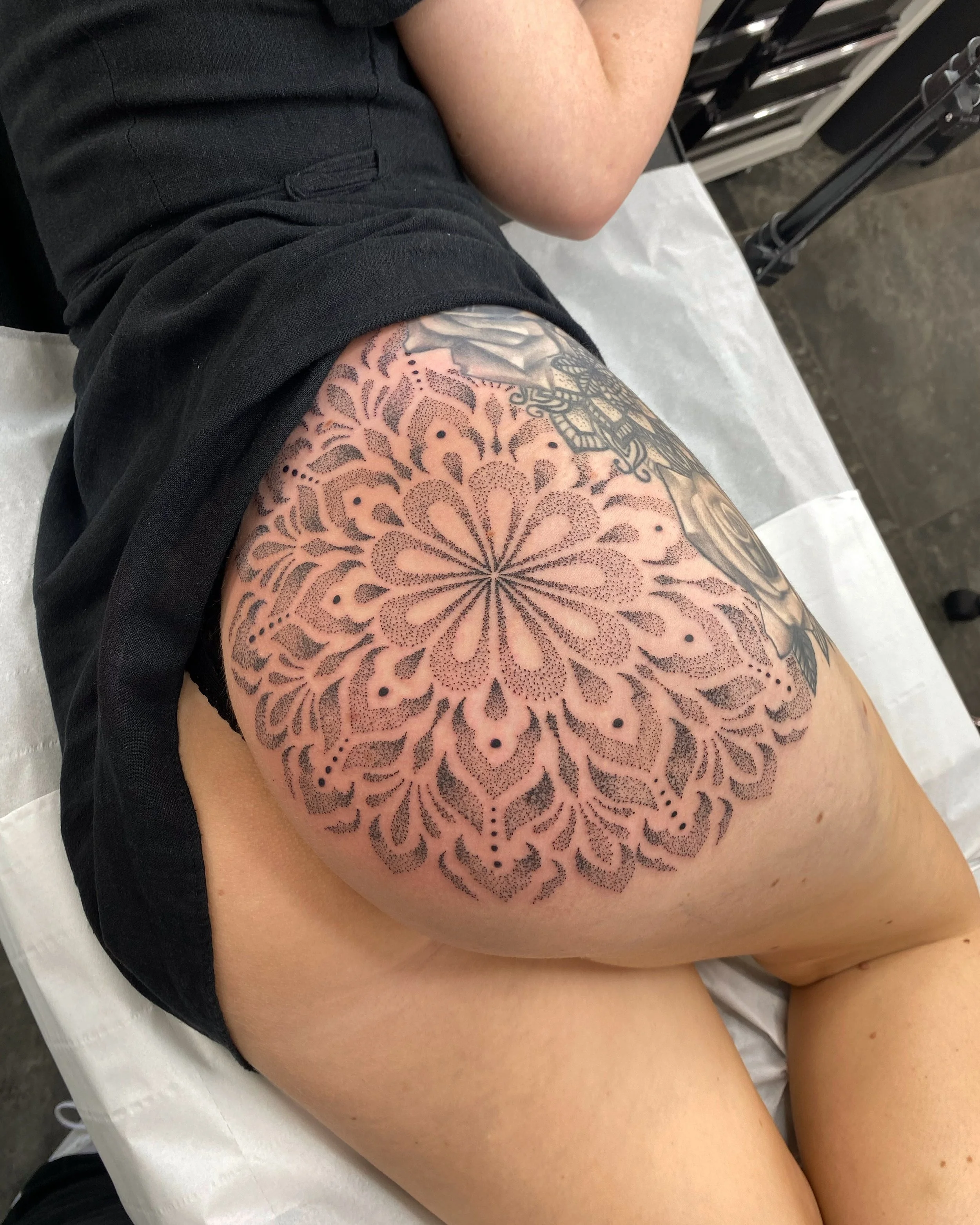 Close-up of a person's upper thigh with a large intricate dotwork tattoo of a mandala or floral pattern.