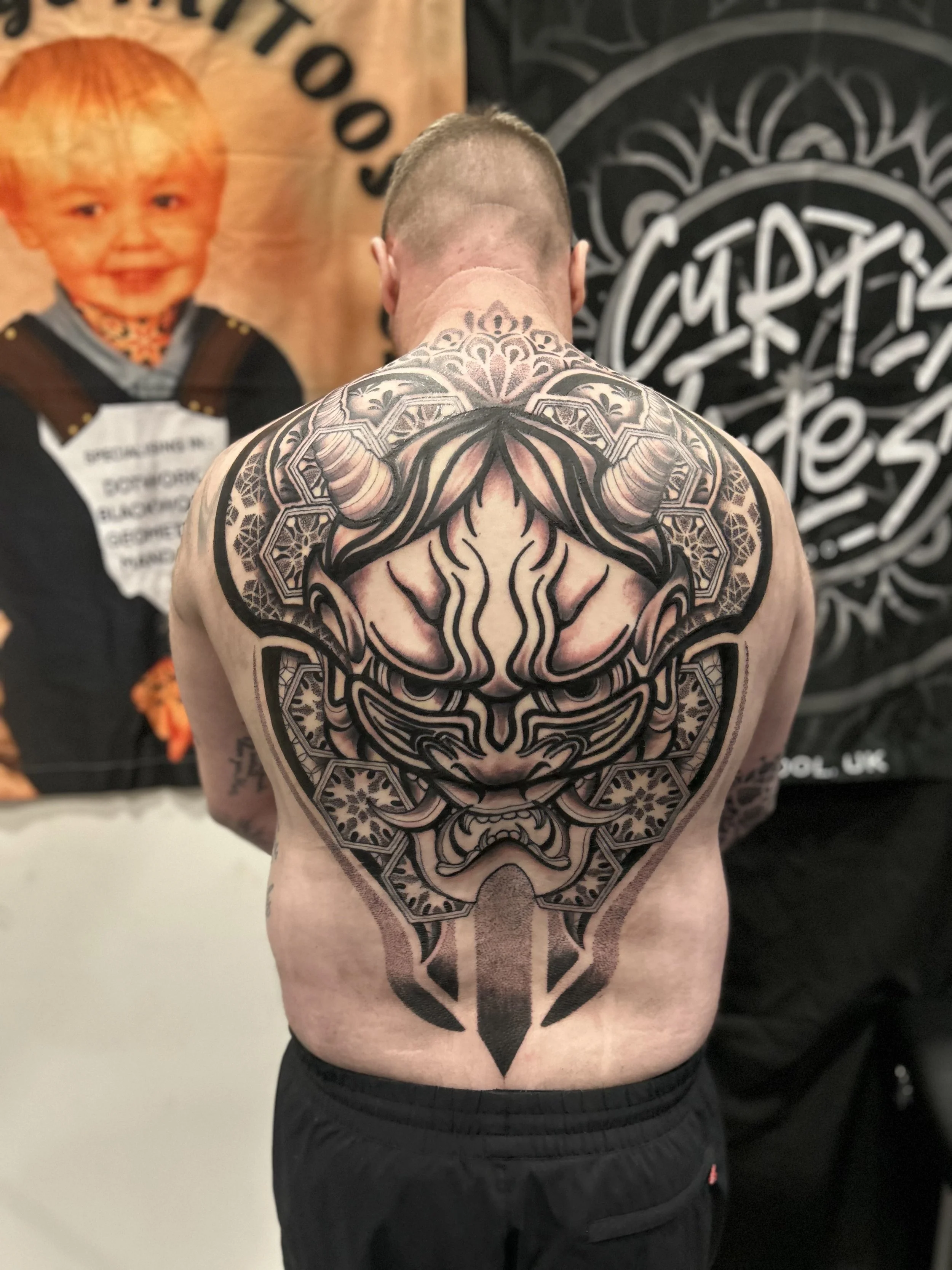 Back of a man with a large, intricate black and gray tattoo covering his shoulders and upper back.