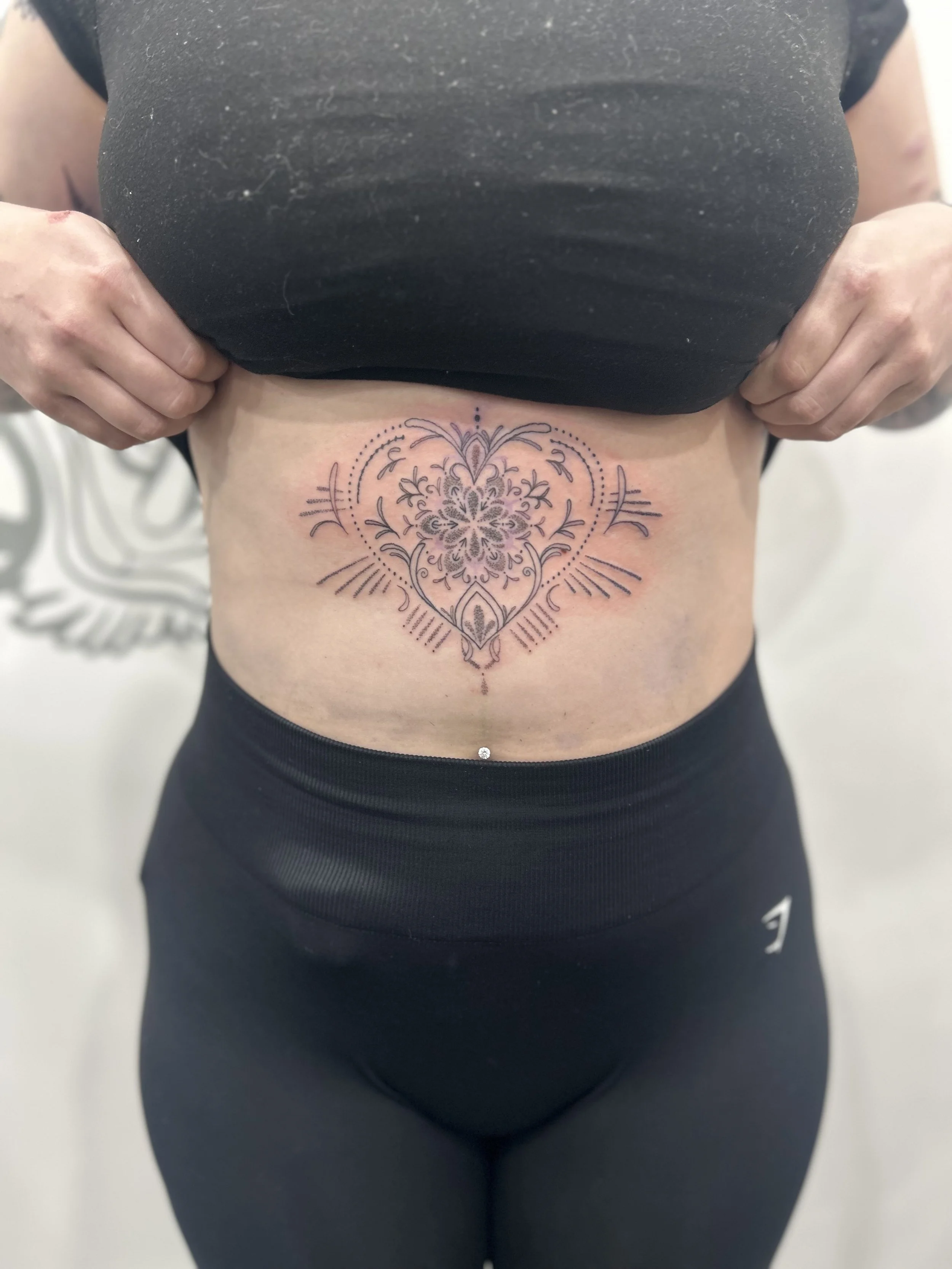 Close-up of a person lifting a black top to reveal a decorative mandala-style tattoo on the lower abdomen, above the waistline of black leggings.