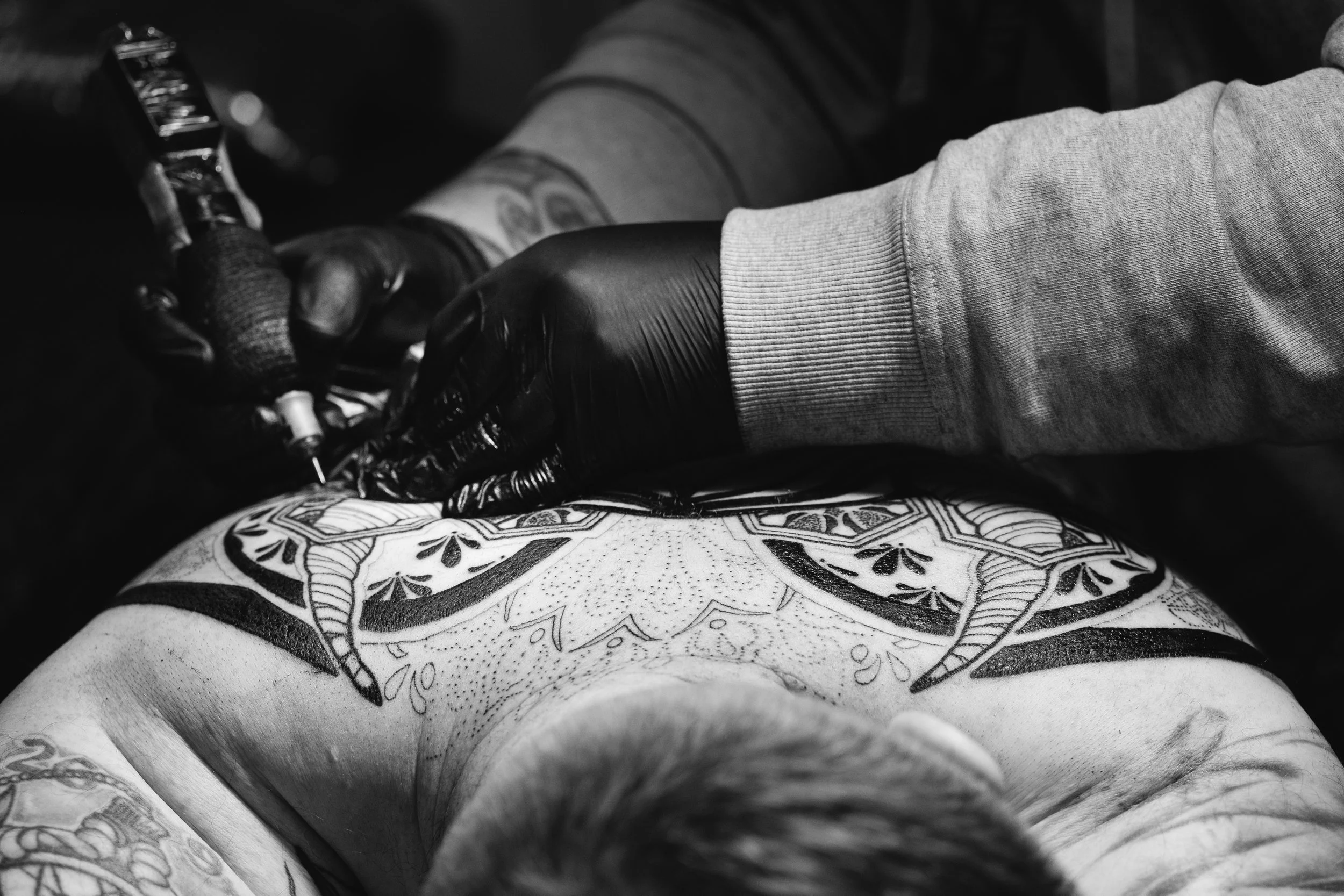 Close-up of a person's tattooed arm and torso, with a tattoo of geometric and floral patterns, as well as a tattoo showing a part of a word or phrase. The person is wearing a gray sweatshirt and is being tattooed by an artist wearing black gloves, using a tattoo machine.