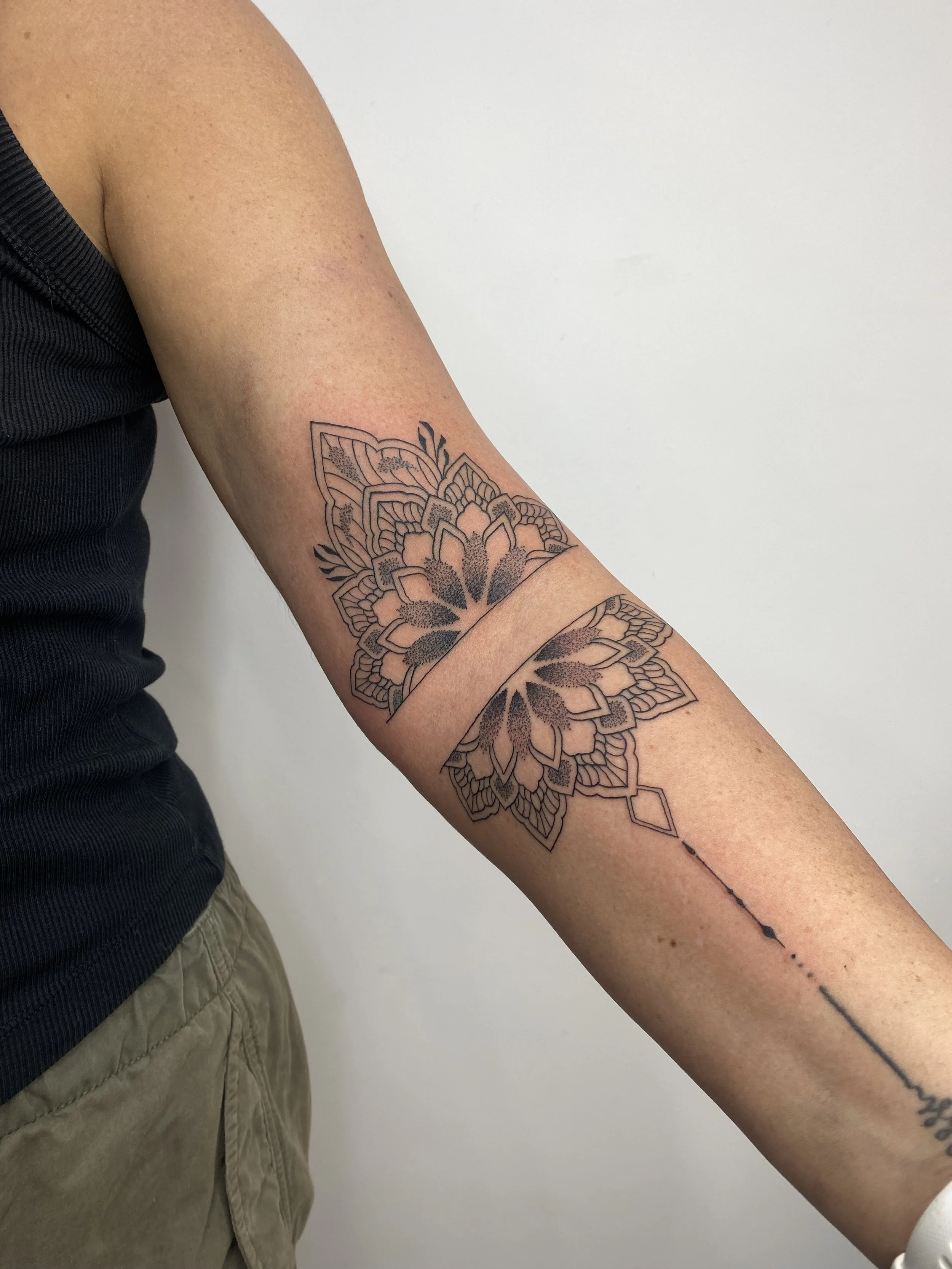 Close-up of a person’s arm with a black and pink floral mandala tattoo and a dotted line tattoo running along the forearm, against a plain white background.